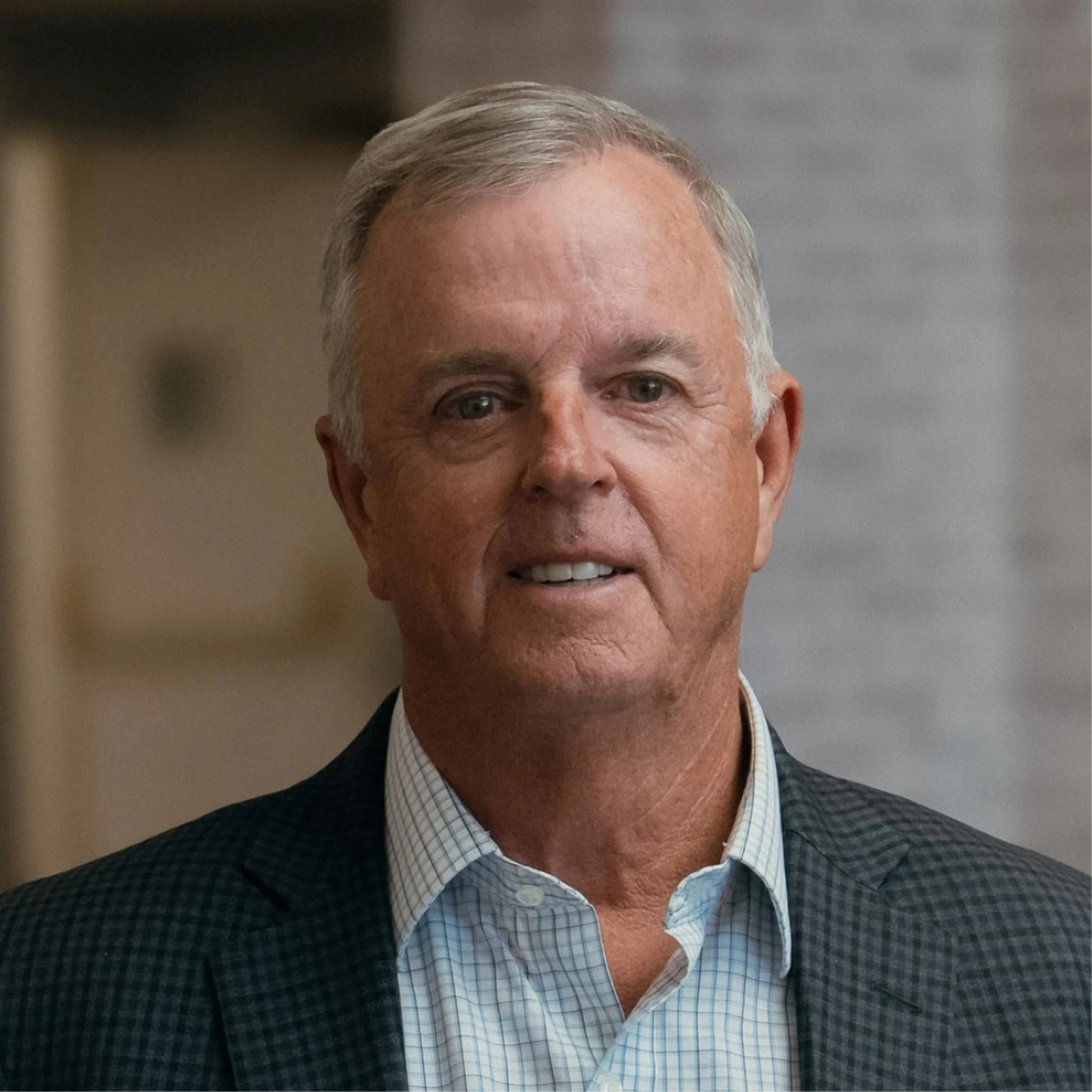 A headshot of a smiling, older white man with short gray hair, wearing a patterned collared shirt and a dark blazer against a blurred indoor background.