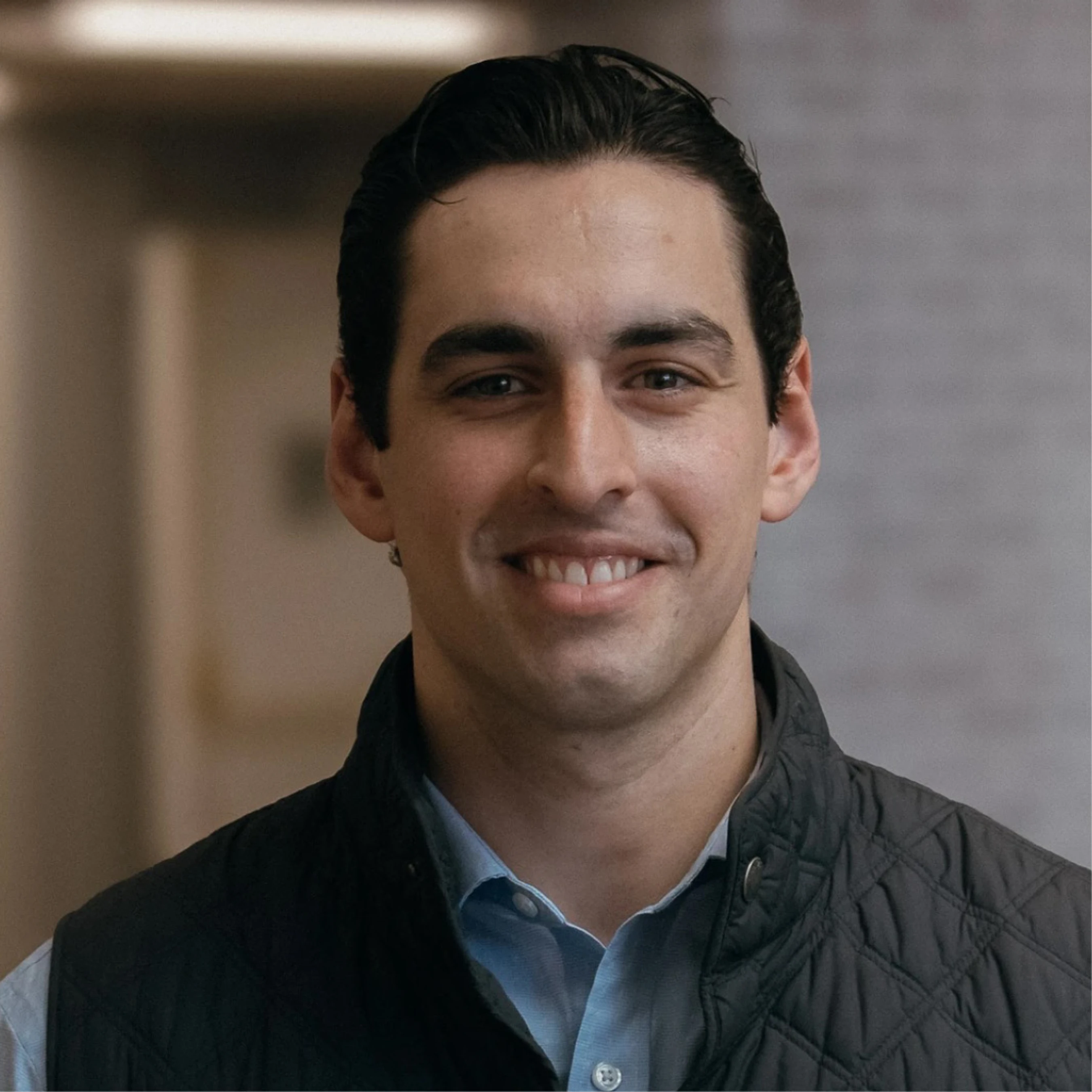 A head-and-shoulders portrait of a smiling young man with dark hair. He is wearing a light blue collared shirt and a dark quilted vest.