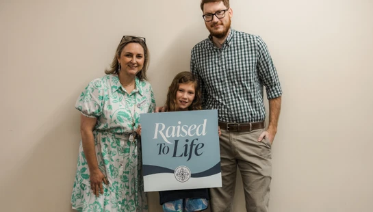 A mother, father, and their young daughter stand smiling for a photo. The girl holds a blue and white sign that reads 'Raised To Life'.