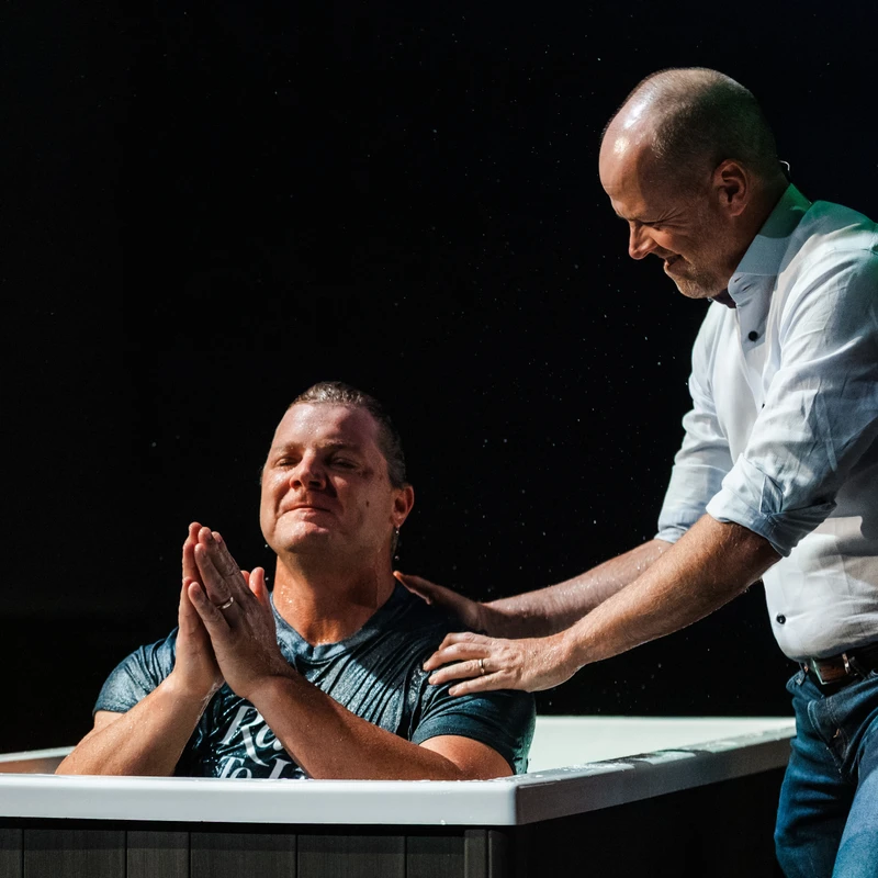 A man sits in a baptismal pool with his hands clasped, while a smiling pastor stands beside him with hands on his shoulders, ready for the baptism.