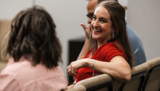 A woman with long brown hair in a red top turns in her seat to smile and laugh with other people in an audience or meeting.
