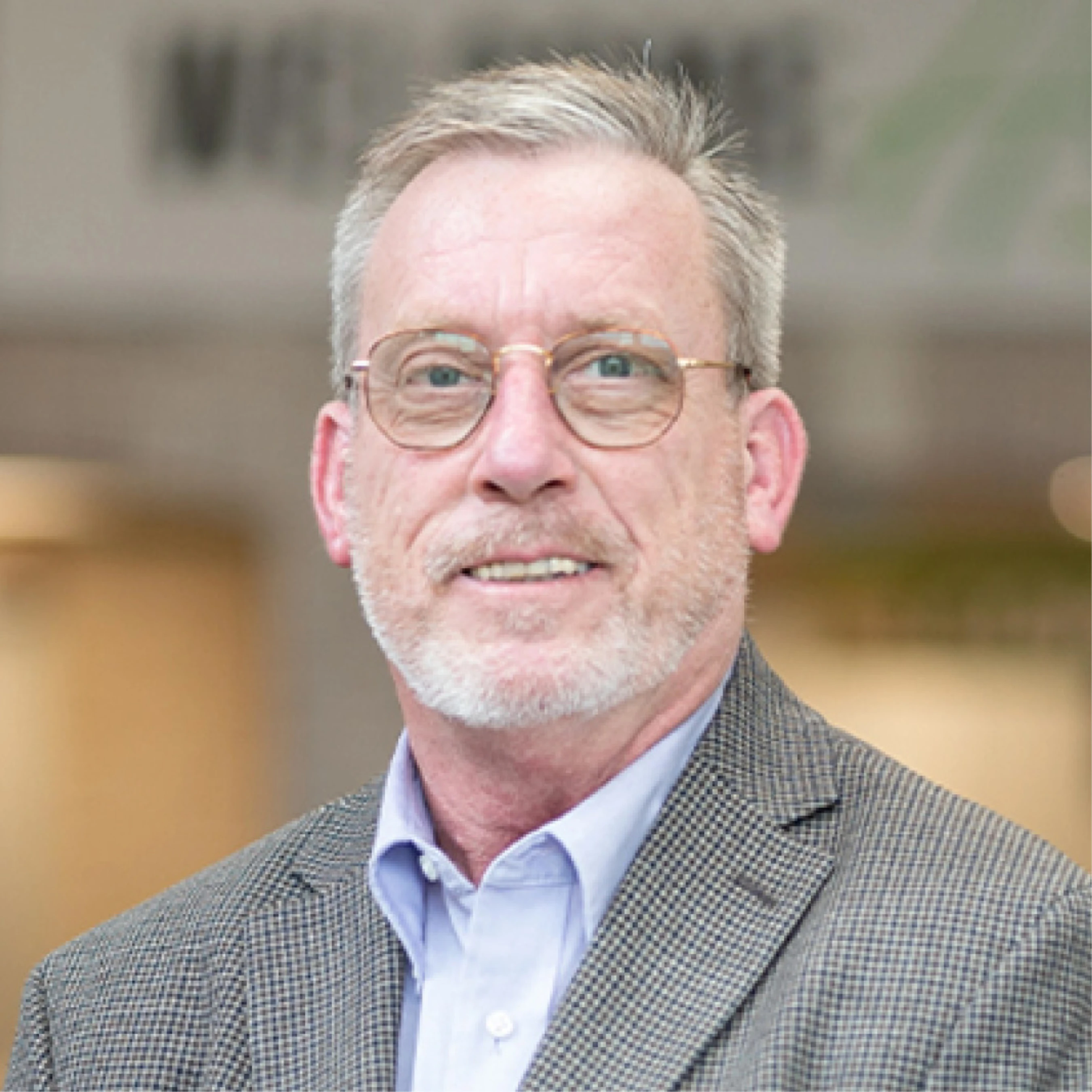 A headshot of a smiling, middle-aged man with graying hair and a short beard. He is wearing glasses, a light blue collared shirt, and a gray blazer.