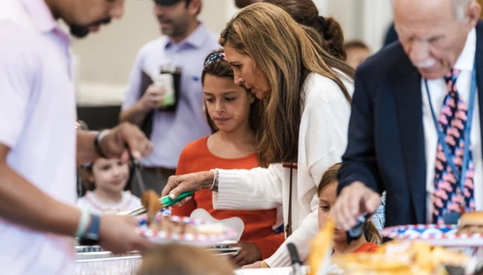 A woman in a white sweater and a young girl stand in a buffet line, serving food onto plates alongside other adults at an indoor community gathering.