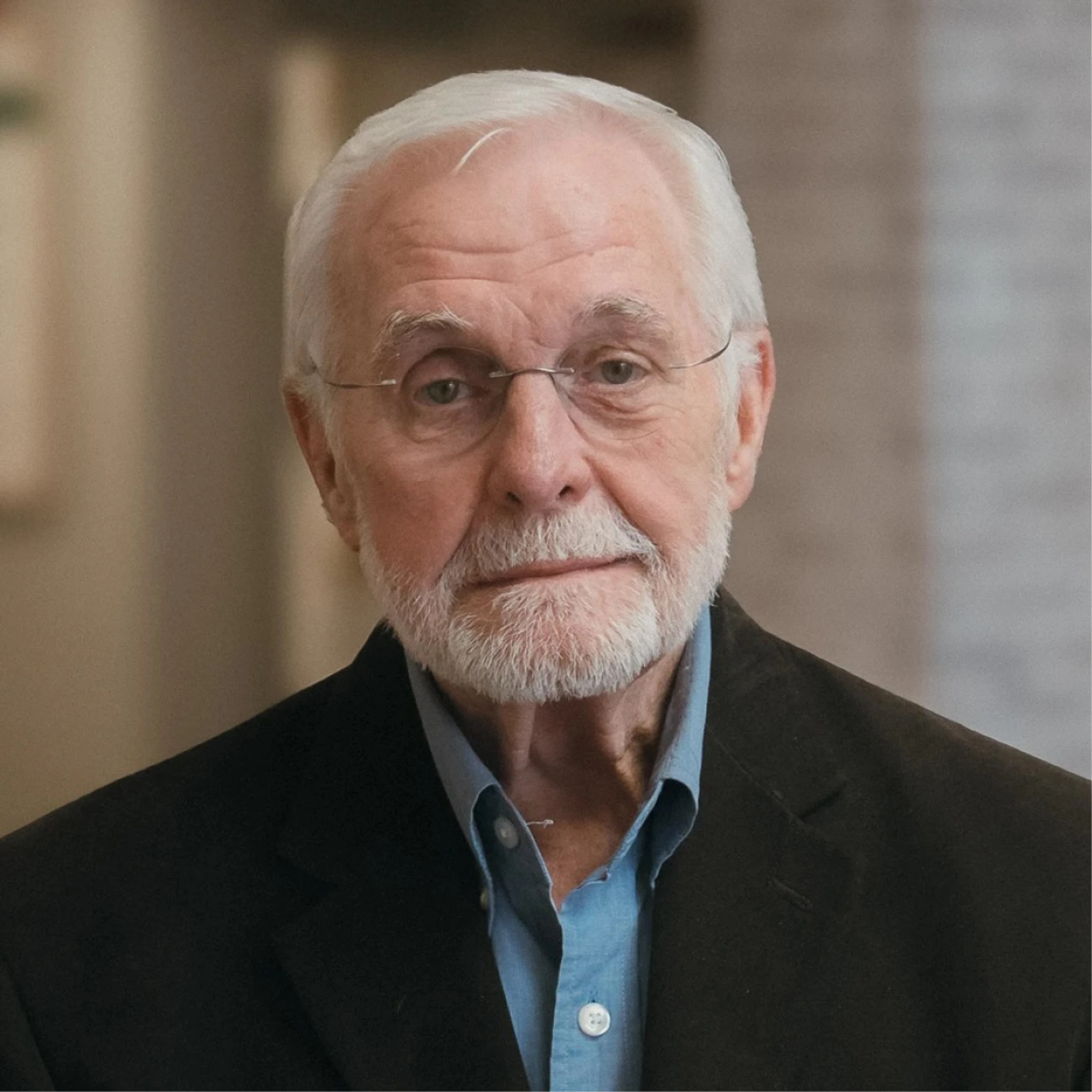 A head-and-shoulders portrait of an elderly man with short white hair, a trimmed white beard, and glasses, wearing a blue shirt and a dark jacket.