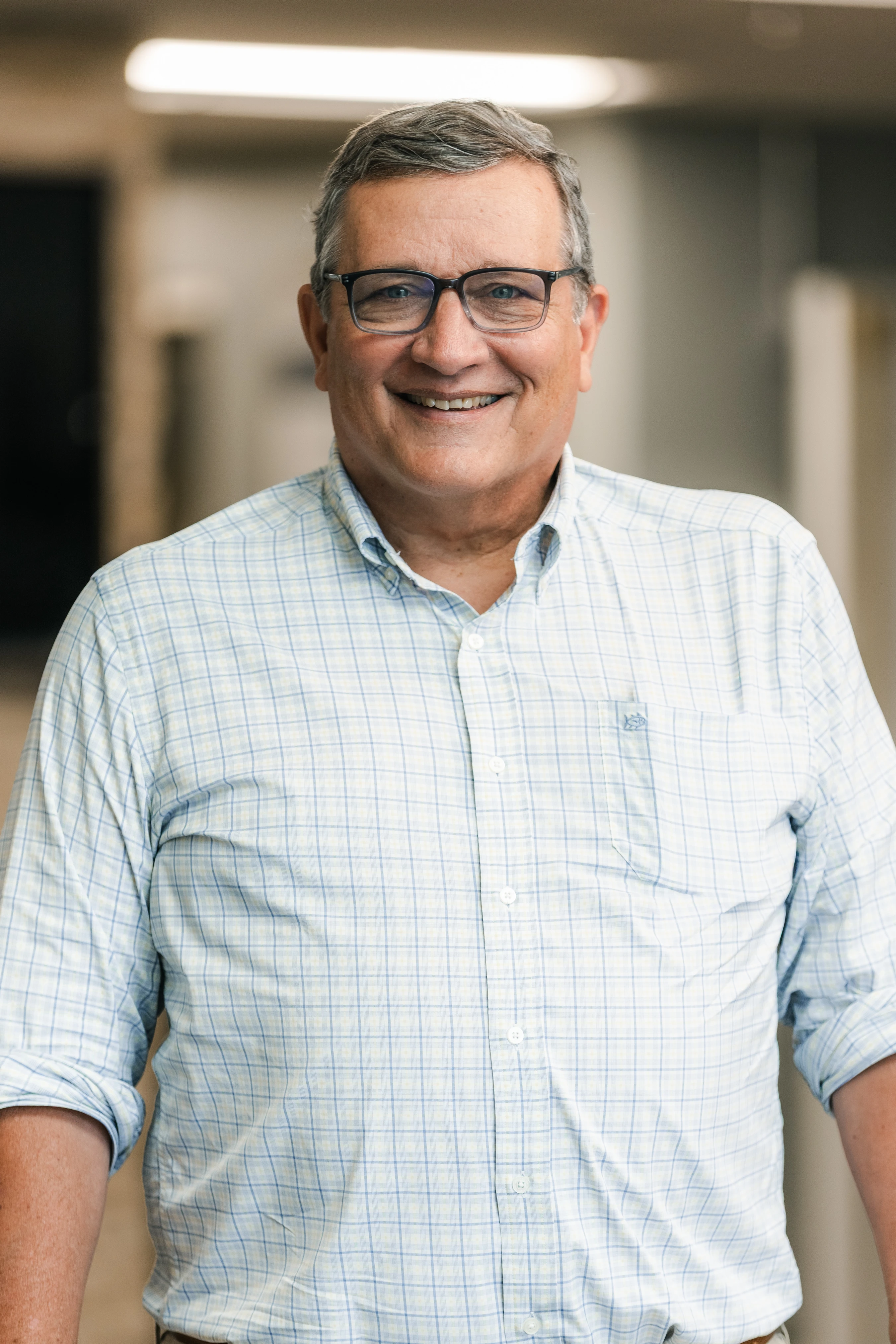 A friendly, older man with gray hair and glasses smiles at the camera. He is wearing a light blue and white plaid button-down shirt in an office hallway.