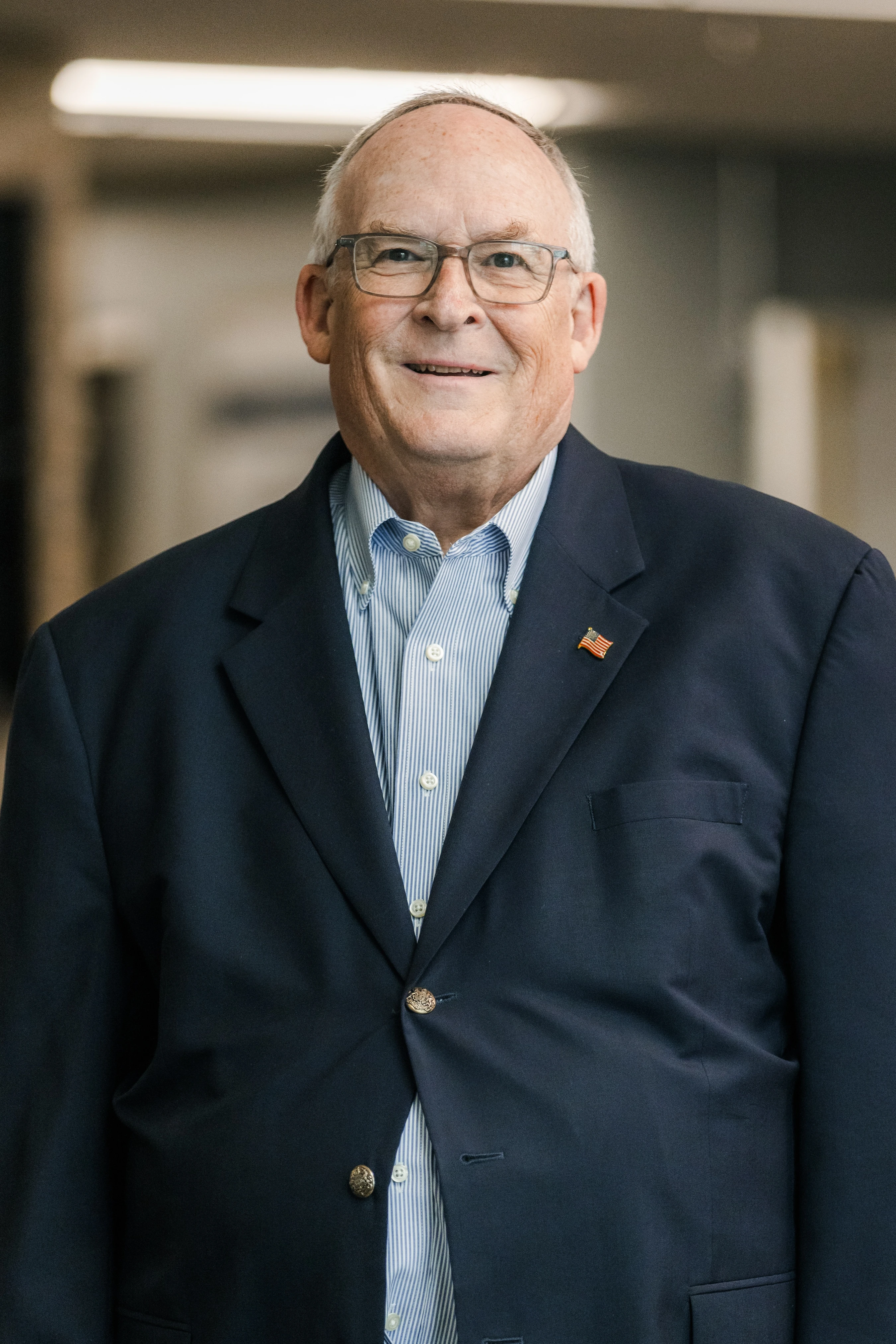 A head-and-shoulders portrait of an older man with glasses, smiling. He wears a navy blazer over a striped button-down shirt and an American flag lapel pin.