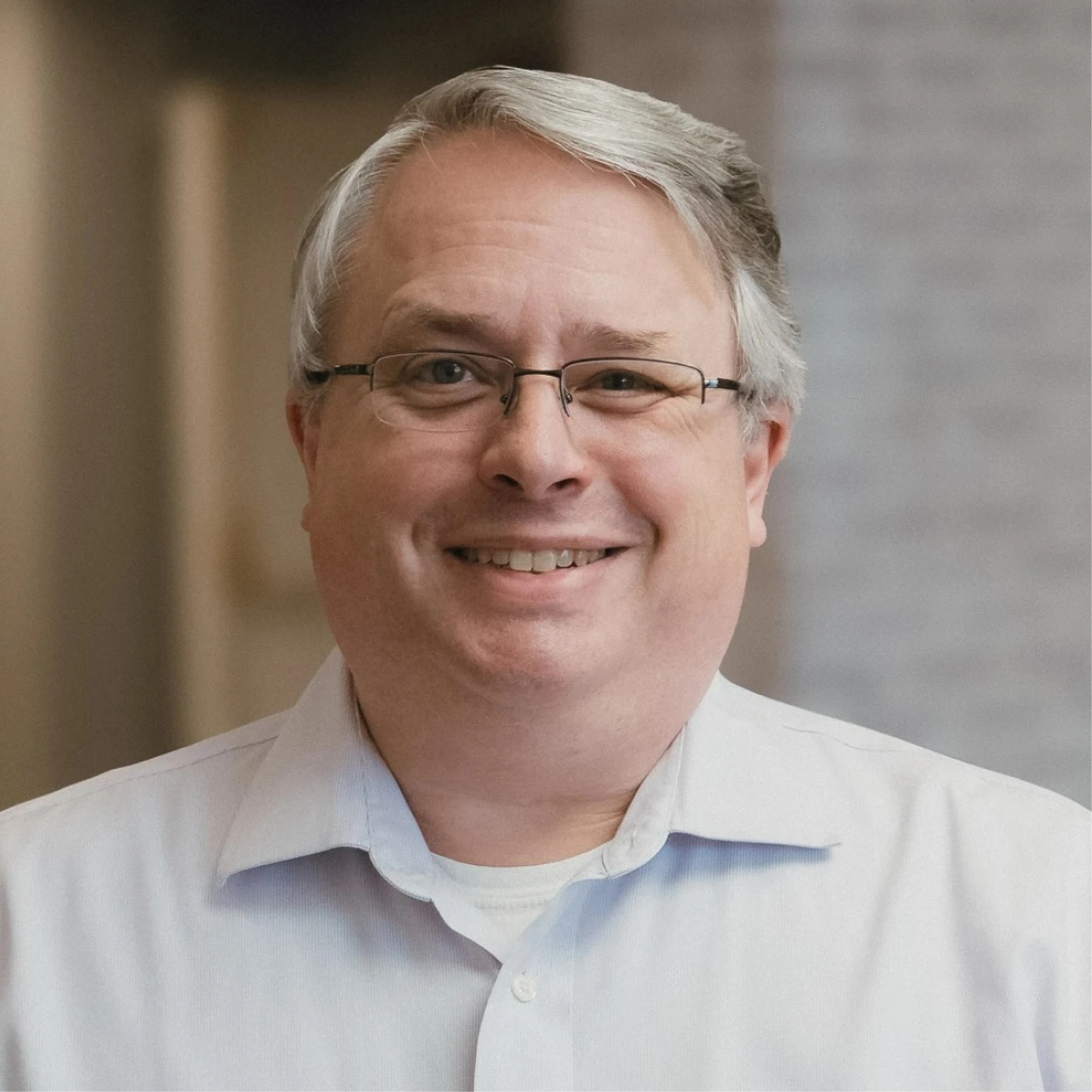 A headshot of a smiling middle-aged man with grey hair and glasses, wearing a light blue collared shirt against a blurred indoor background.