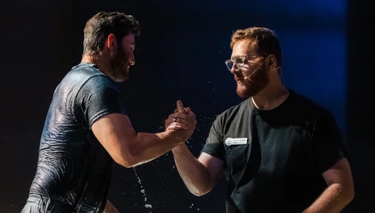 A wet man in a dark T-shirt joyfully clasps hands with a pastor in glasses after being baptized. Water drips from his arms against a dark blue background.