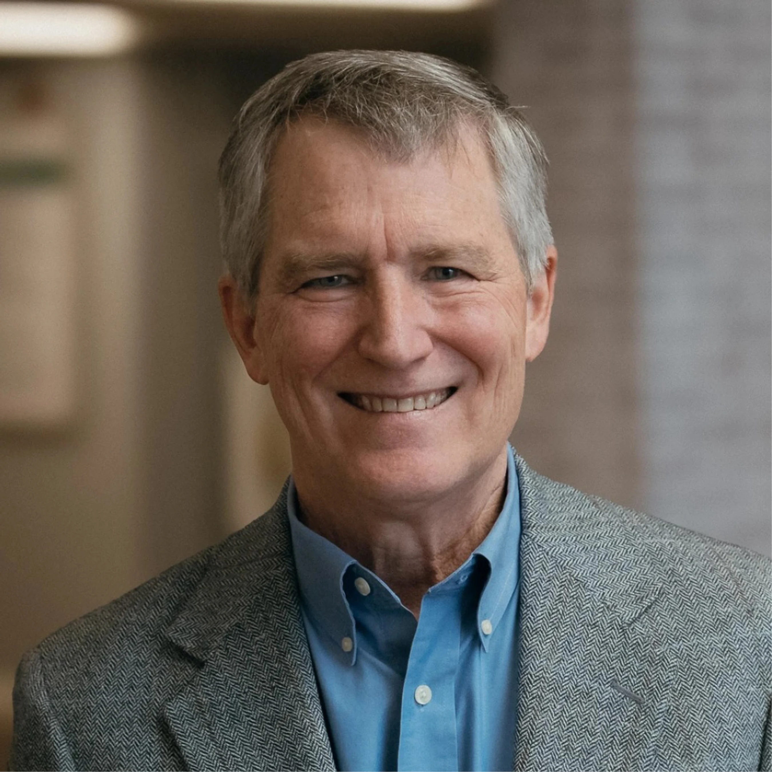 A headshot of a smiling older man with grey hair and blue eyes. He is wearing a blue button-down shirt and a grey herringbone blazer.