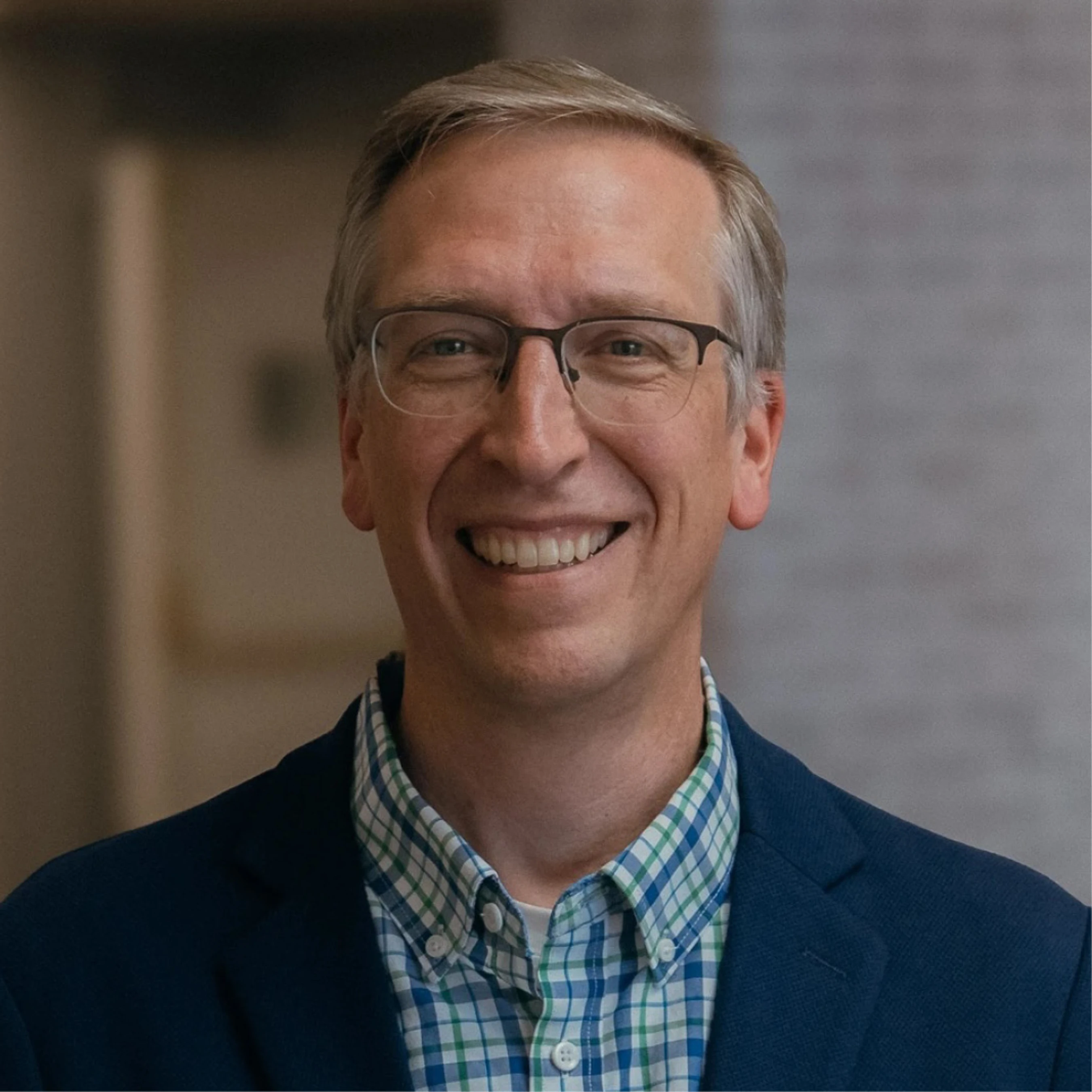 A professional headshot of a middle-aged man with short graying hair and glasses, smiling at the camera. He wears a checked shirt and a blue blazer.