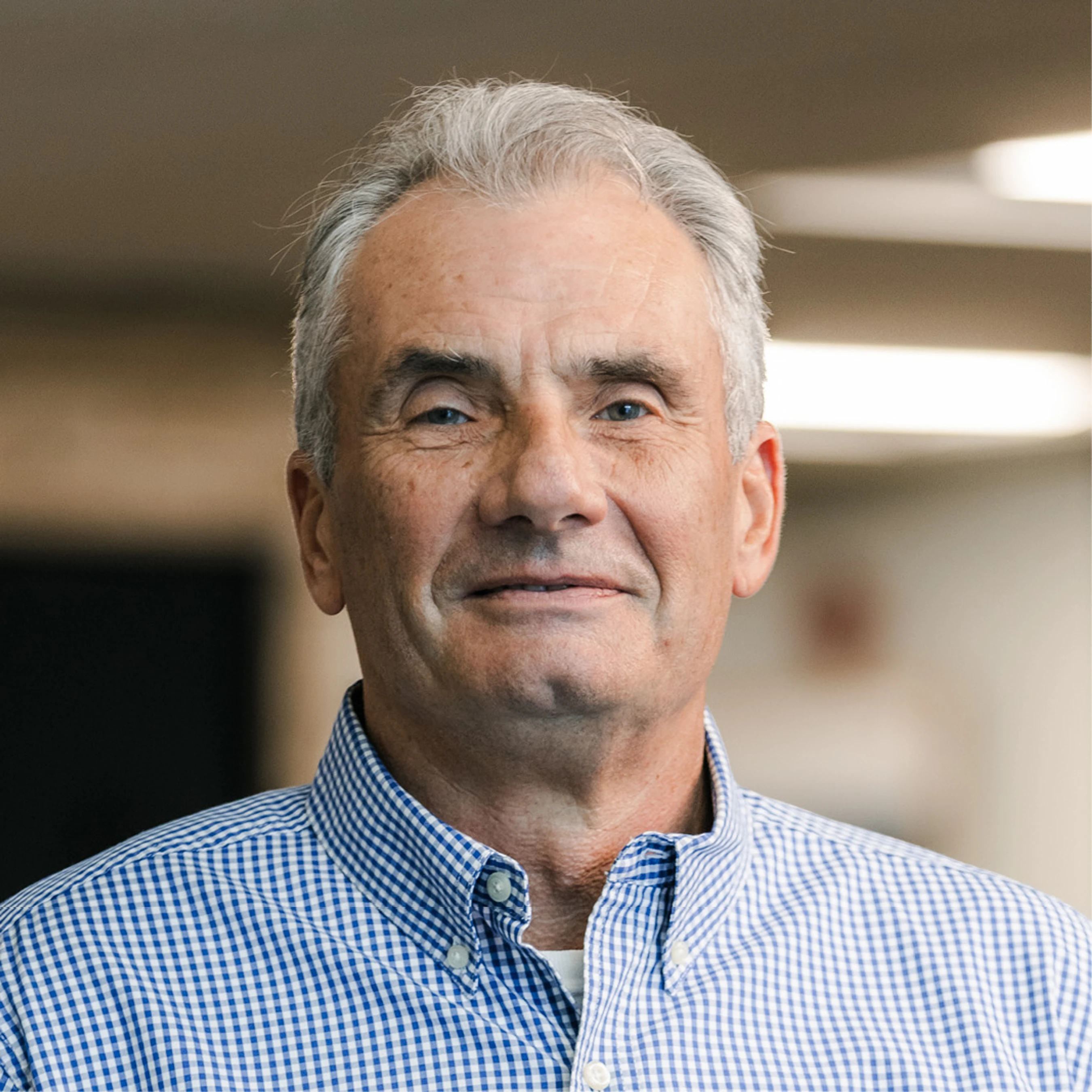 A professional headshot of an older man with gray hair and blue eyes, smiling slightly. He wears a blue and white checkered button-down shirt.