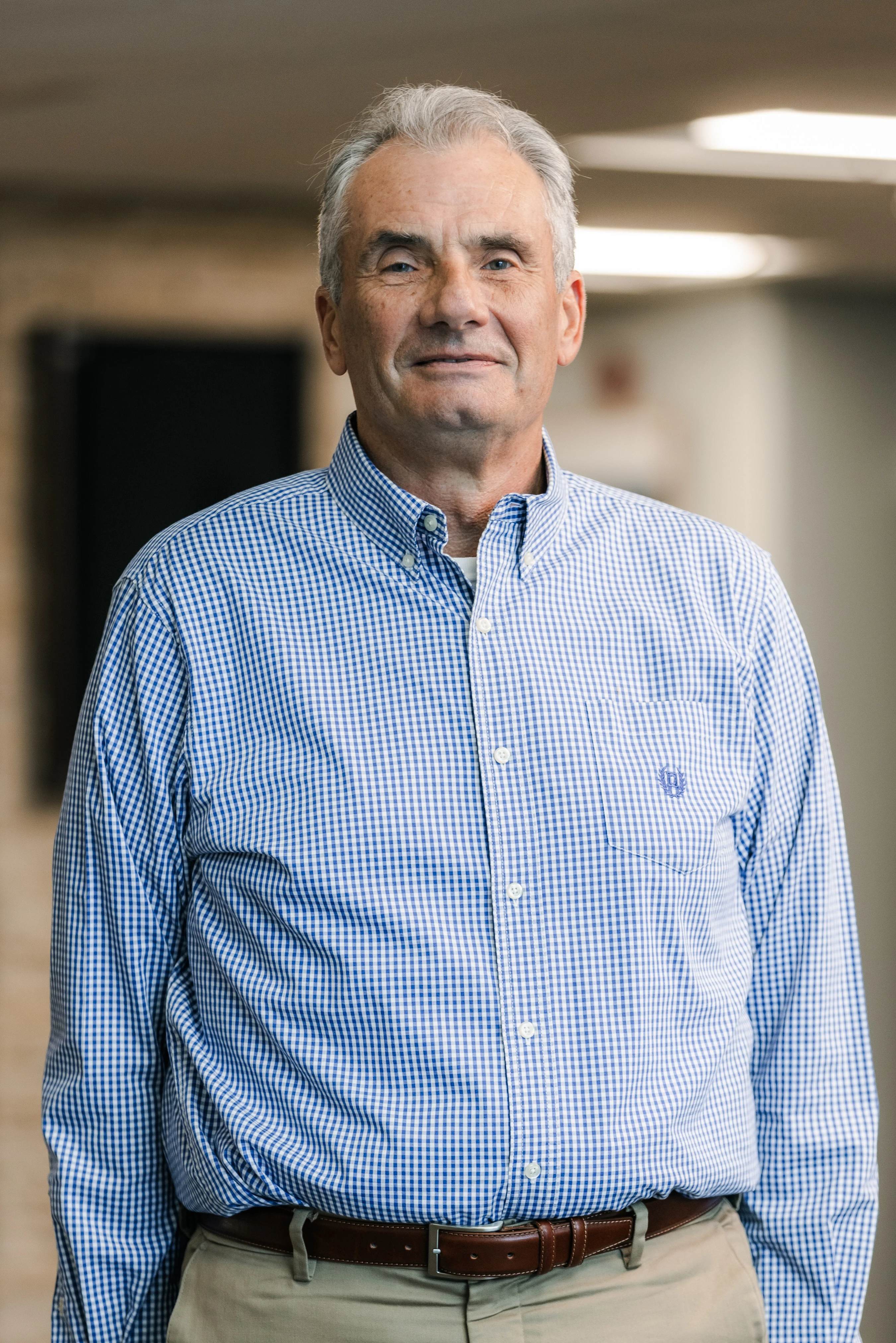 A man with gray hair and a pleasant expression poses for a portrait in a hallway. He is wearing a blue and white checkered button-down shirt and khaki pants.
