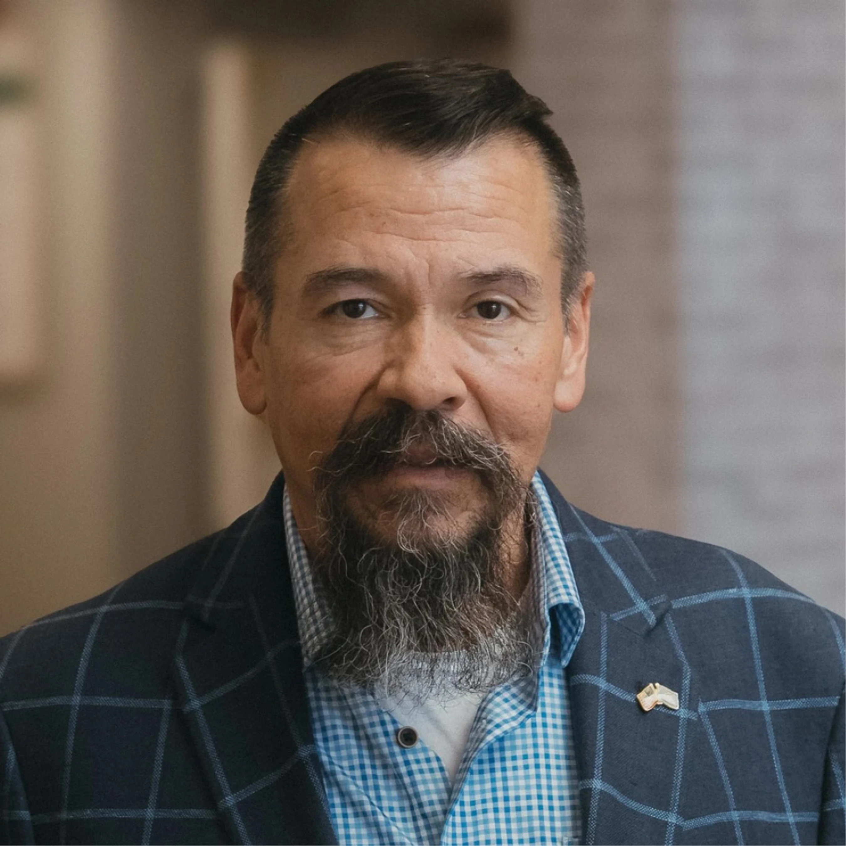 A headshot of a middle-aged man with a mustache and goatee, wearing a blue plaid blazer over a checkered shirt. He looks seriously at the camera.