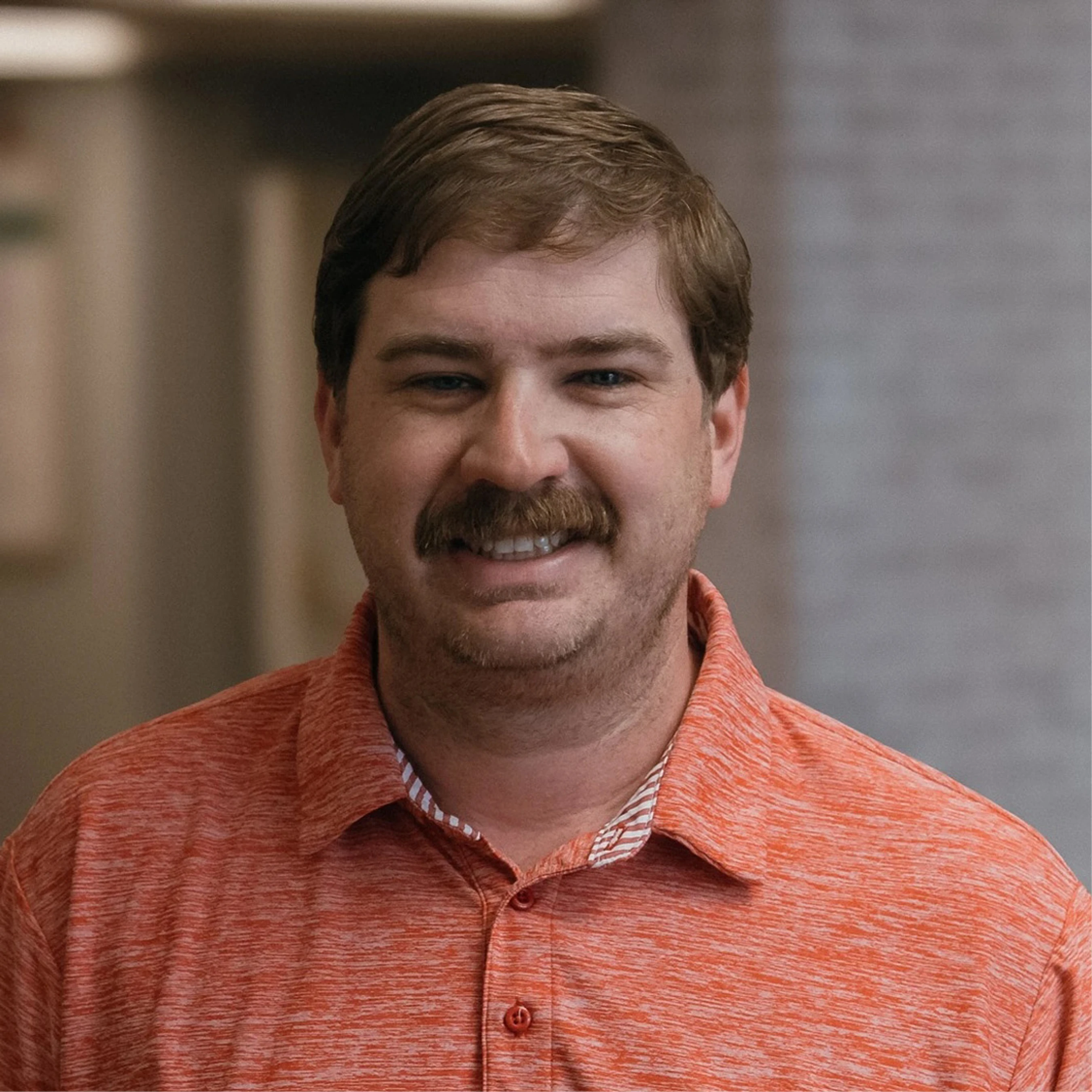 A headshot of a caucasian man with brown hair and a mustache, smiling at the camera. He is wearing an orange collared polo shirt in an office setting.