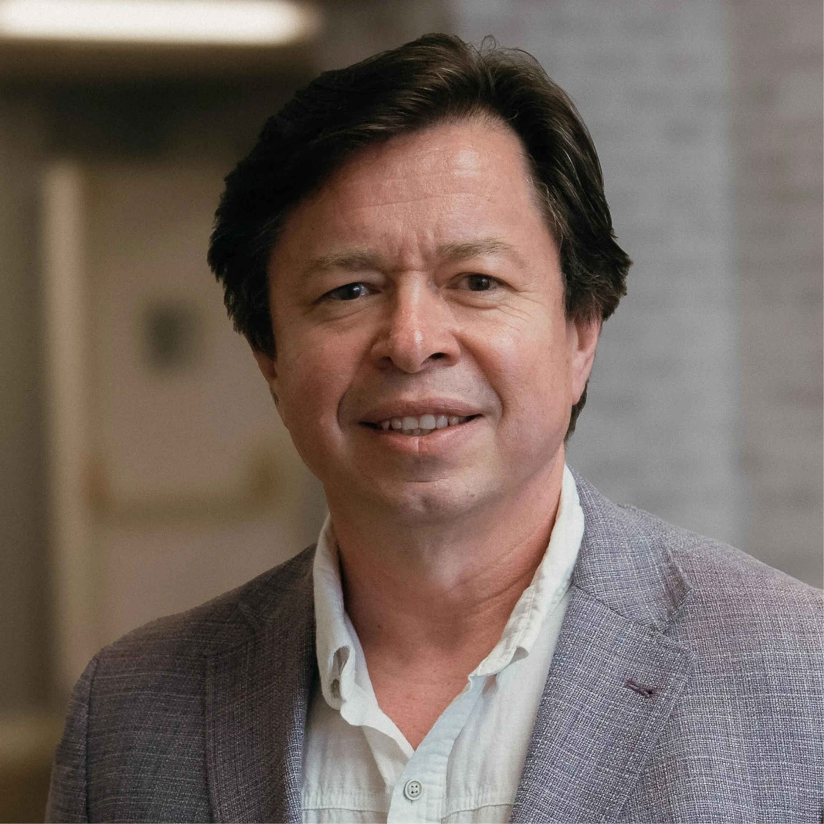 A headshot of a smiling, middle-aged man with dark hair. He is wearing a light gray blazer over a white button-down shirt, against a blurred background.