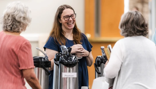 A woman with glasses and long brown hair laughs while talking with two other women at a coffee station with large silver urns.