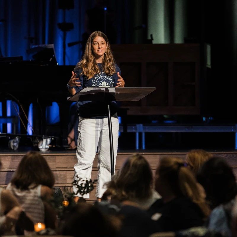 A woman with long brown hair stands on a wooden stage behind a lectern, speaking to an audience. She wears a dark t-shirt and white pants in a dimly lit hall.