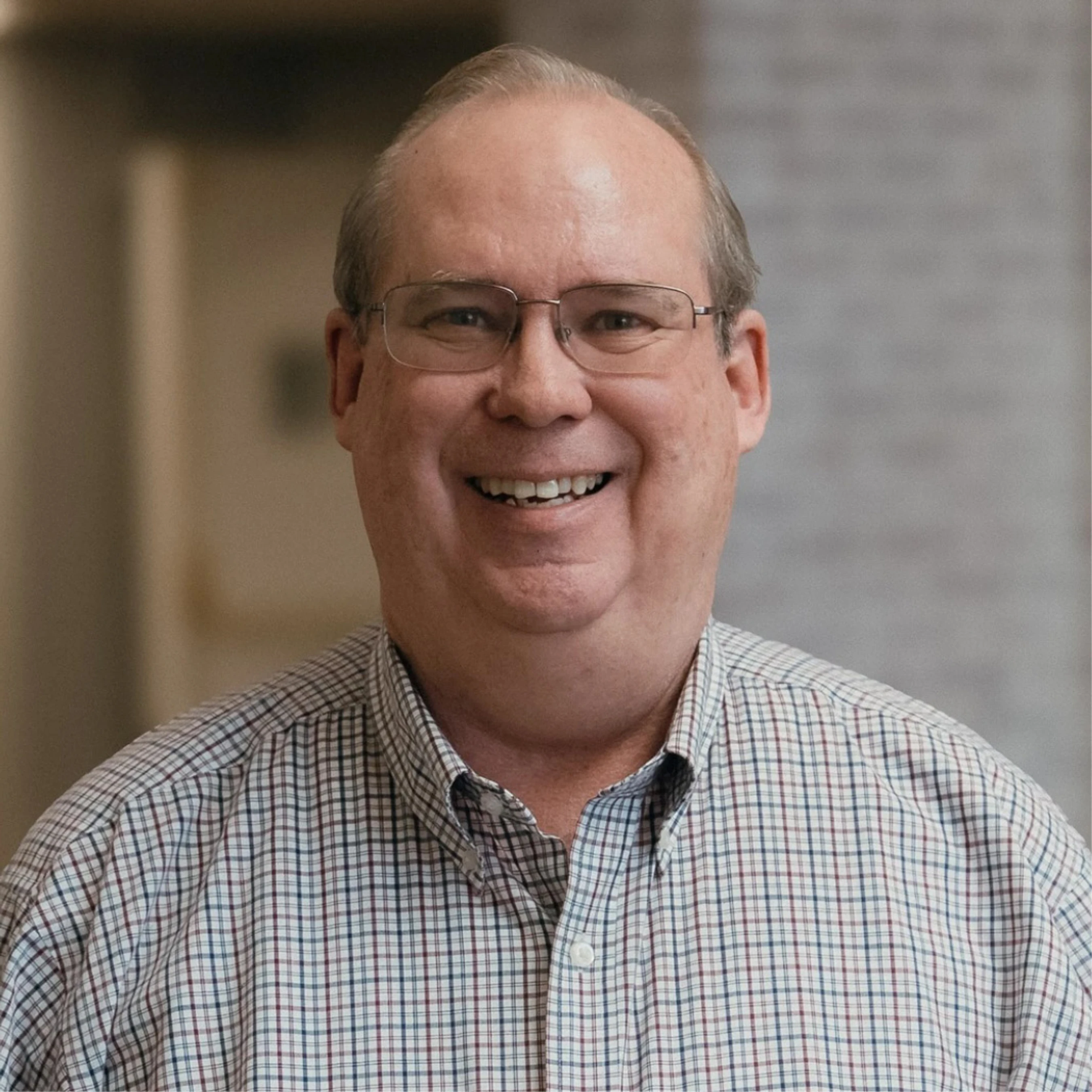 A friendly, middle-aged man with glasses and thinning hair smiles for a headshot. He is wearing a plaid button-down shirt against a blurred indoor background.