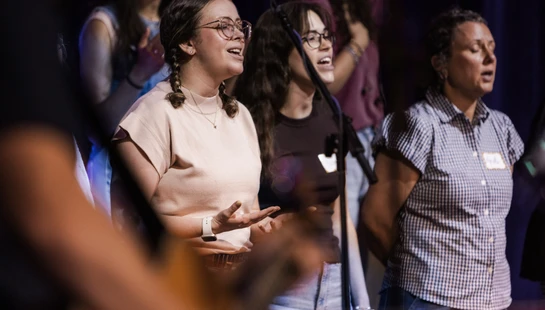 Three women stand side-by-side singing. The central woman wears glasses and has braids, singing with open hands. The stage is dark behind them.