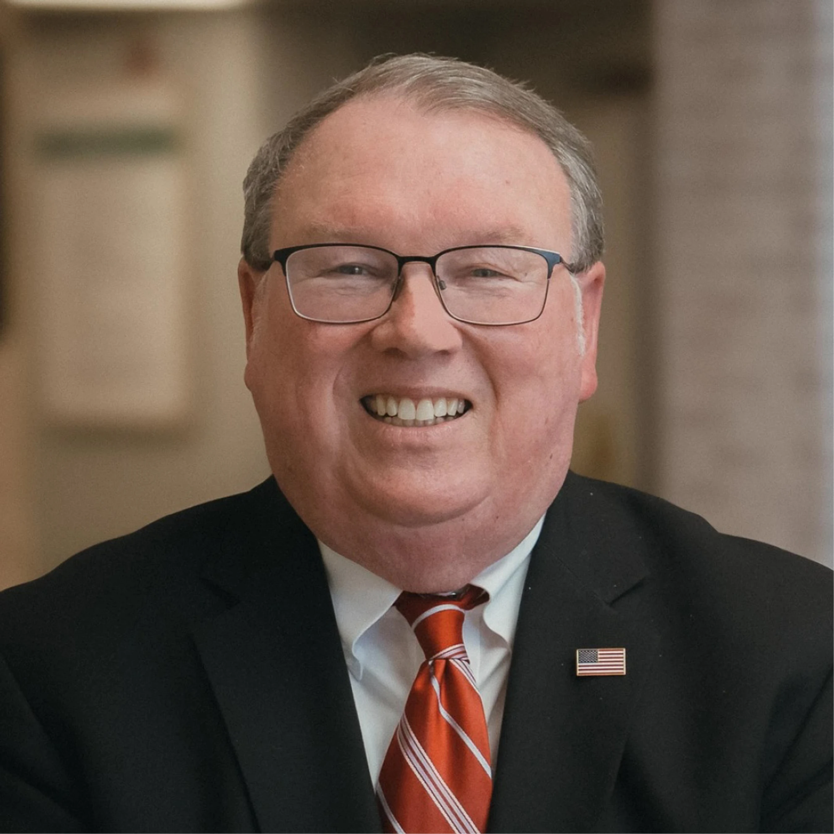 A headshot of a smiling, middle-aged man with glasses. He is wearing a black suit jacket, white shirt, red striped tie, and an American flag lapel pin.