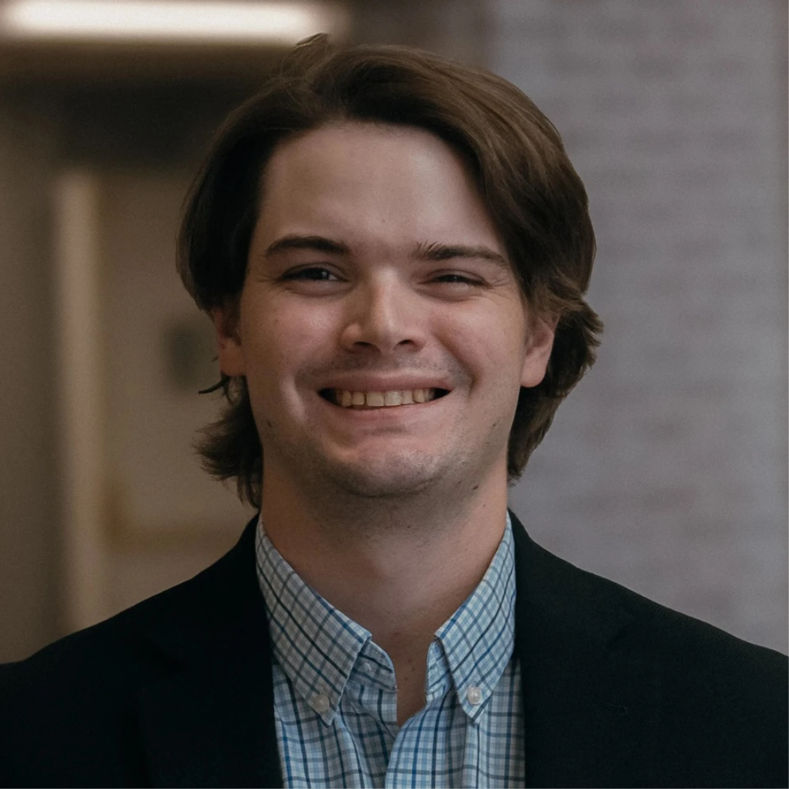A headshot of a smiling young man with medium-length brown hair, wearing a blue checkered button-down shirt and a dark blazer.