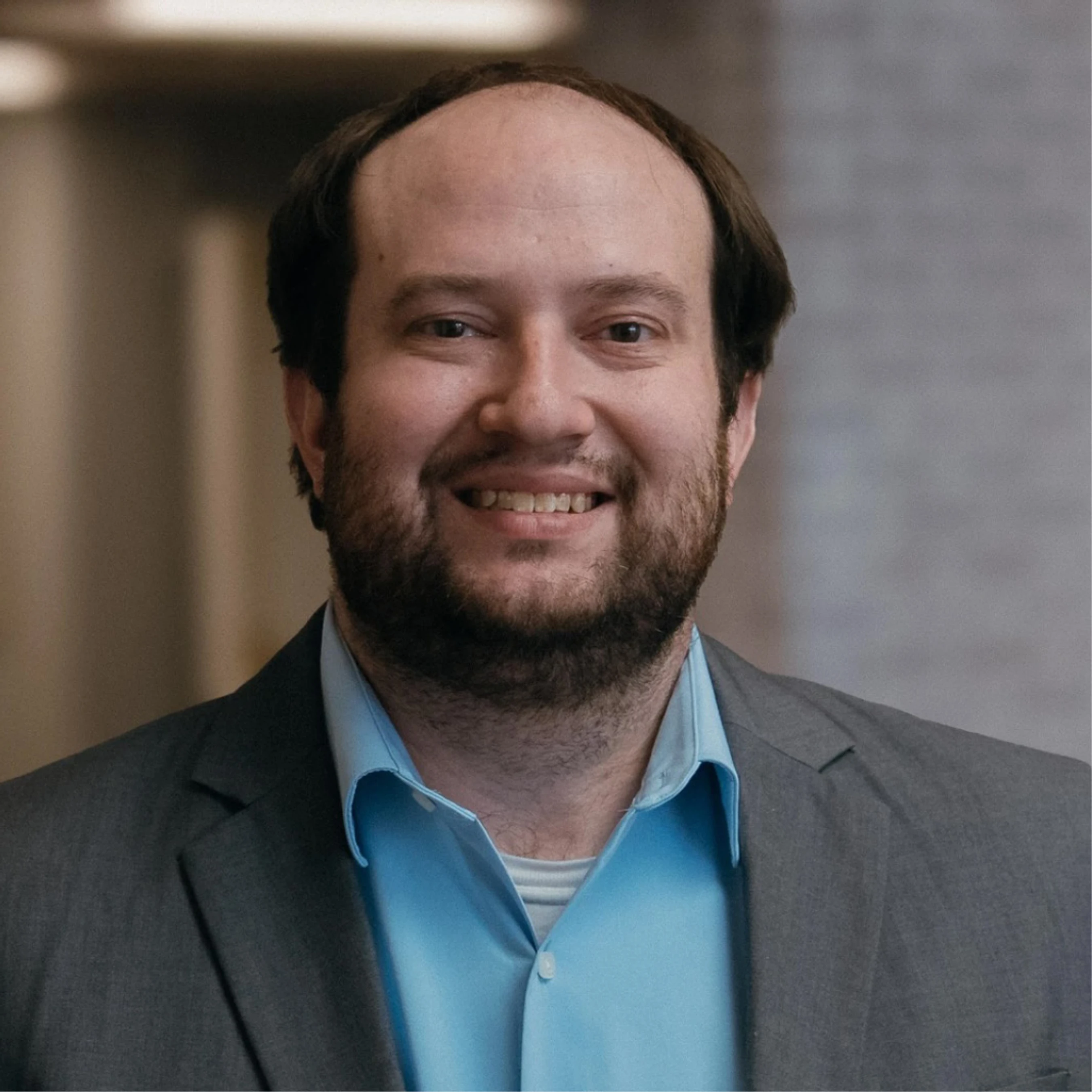 A head and shoulders portrait of a smiling man with receding brown hair and a beard, wearing a light blue shirt and a grey suit jacket in an office setting.