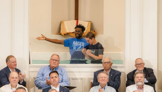 A smiling pastor baptizes a young man in a church baptismal font while a congregation of men seated in front of them applauds.