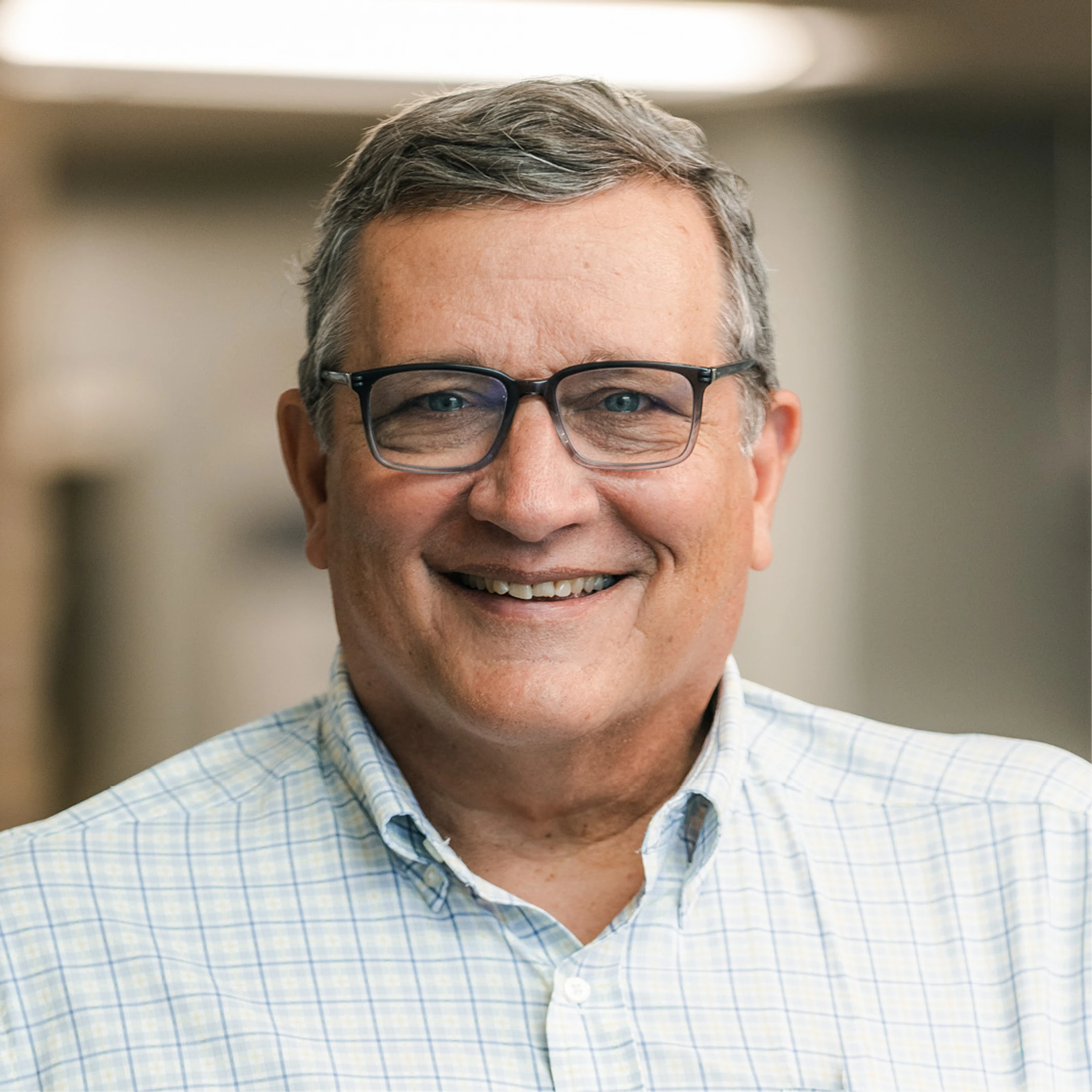 A professional headshot of a smiling, middle-aged man with graying hair and glasses, wearing a light-colored plaid button-down shirt.
