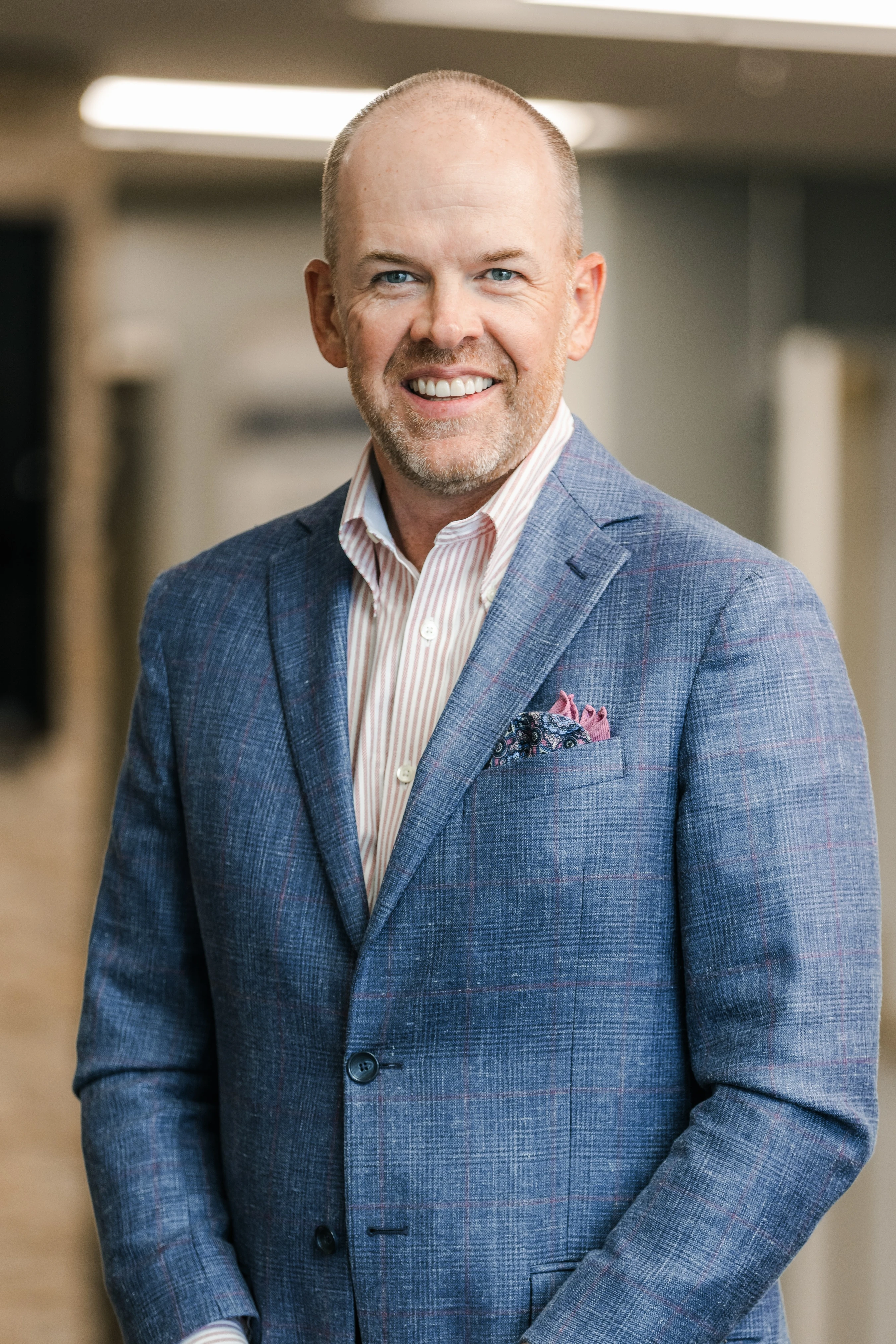 A head and shoulders portrait of a smiling man with blue eyes and short hair. He is wearing a blue plaid sport coat, a striped shirt, and a pocket square.