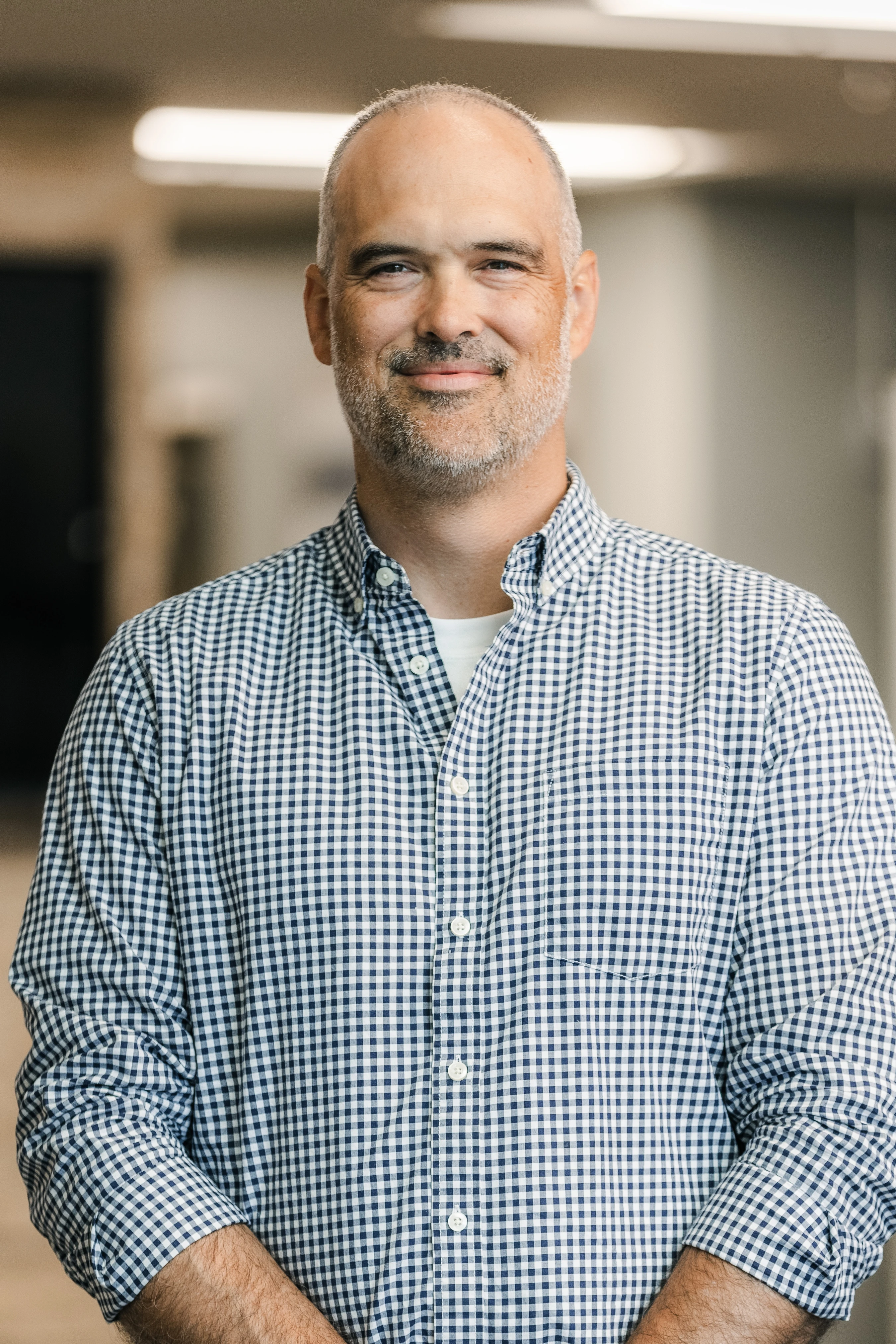 A headshot of a middle-aged man with short graying hair and a beard, smiling warmly while wearing a blue and white checkered button-down shirt.