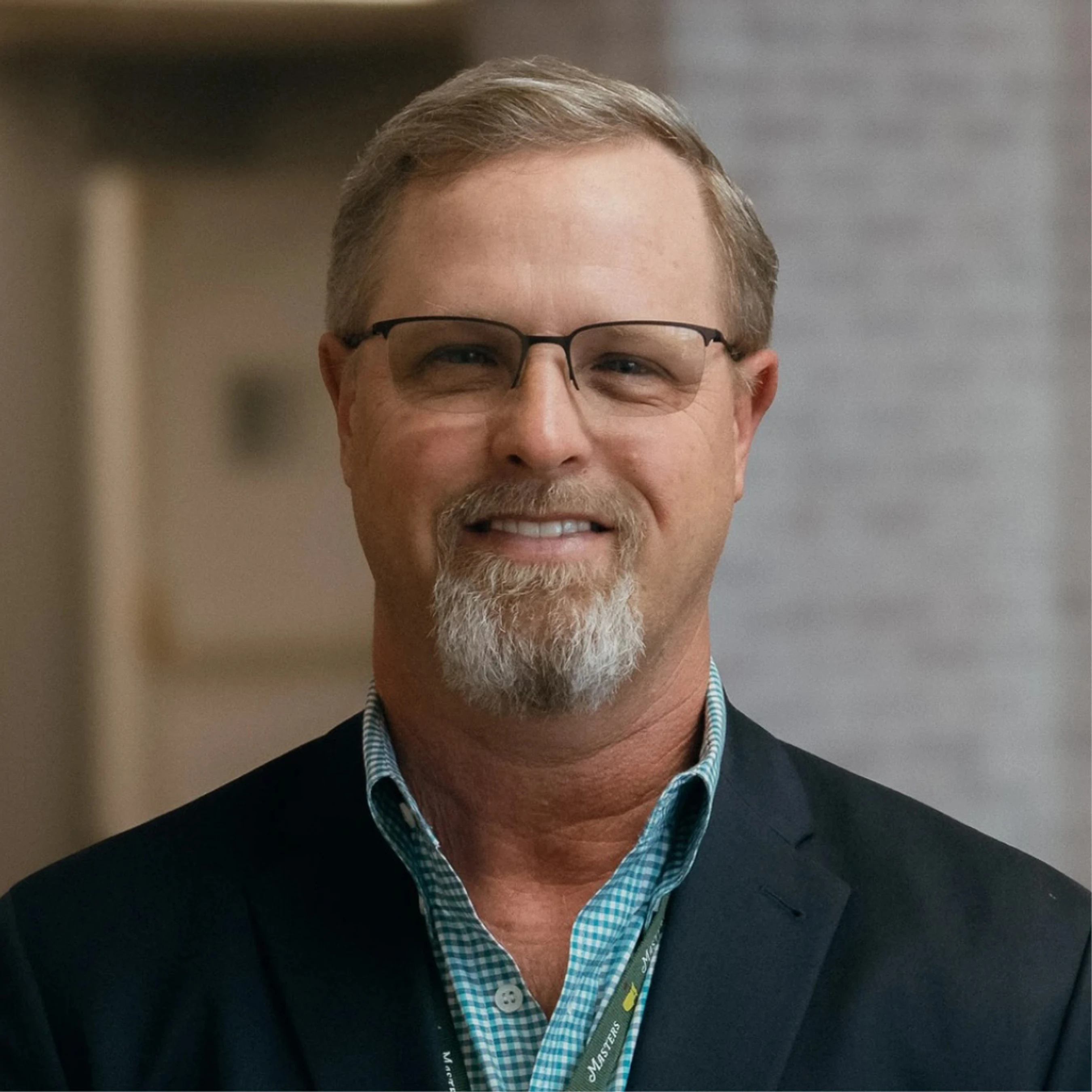 A head and shoulders portrait of a smiling middle-aged man with glasses and a gray goatee, wearing a checkered shirt and a dark blazer indoors.