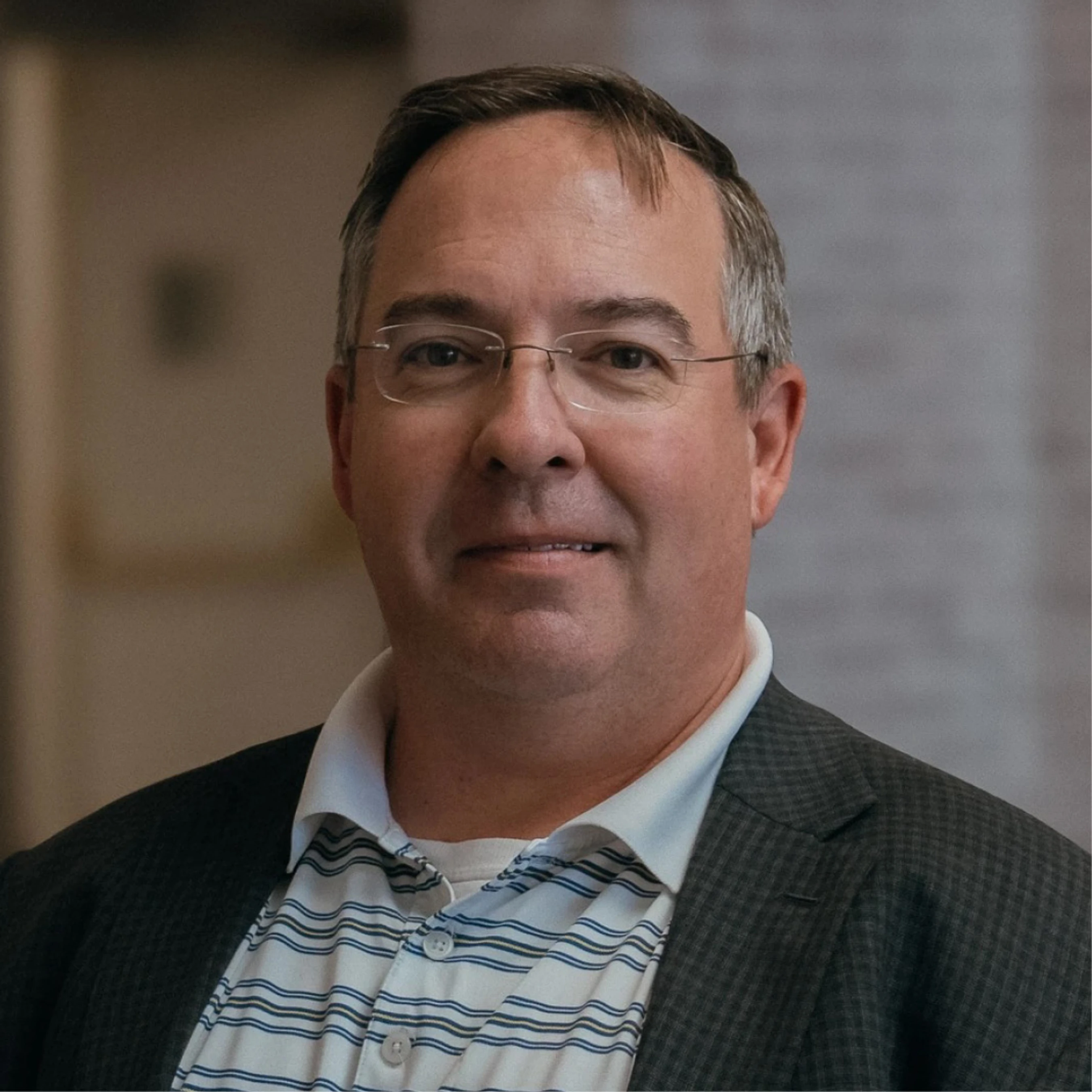 A head-and-shoulders portrait of a middle-aged man with glasses, wearing a dark blazer over a striped polo shirt, smiling slightly at the camera.