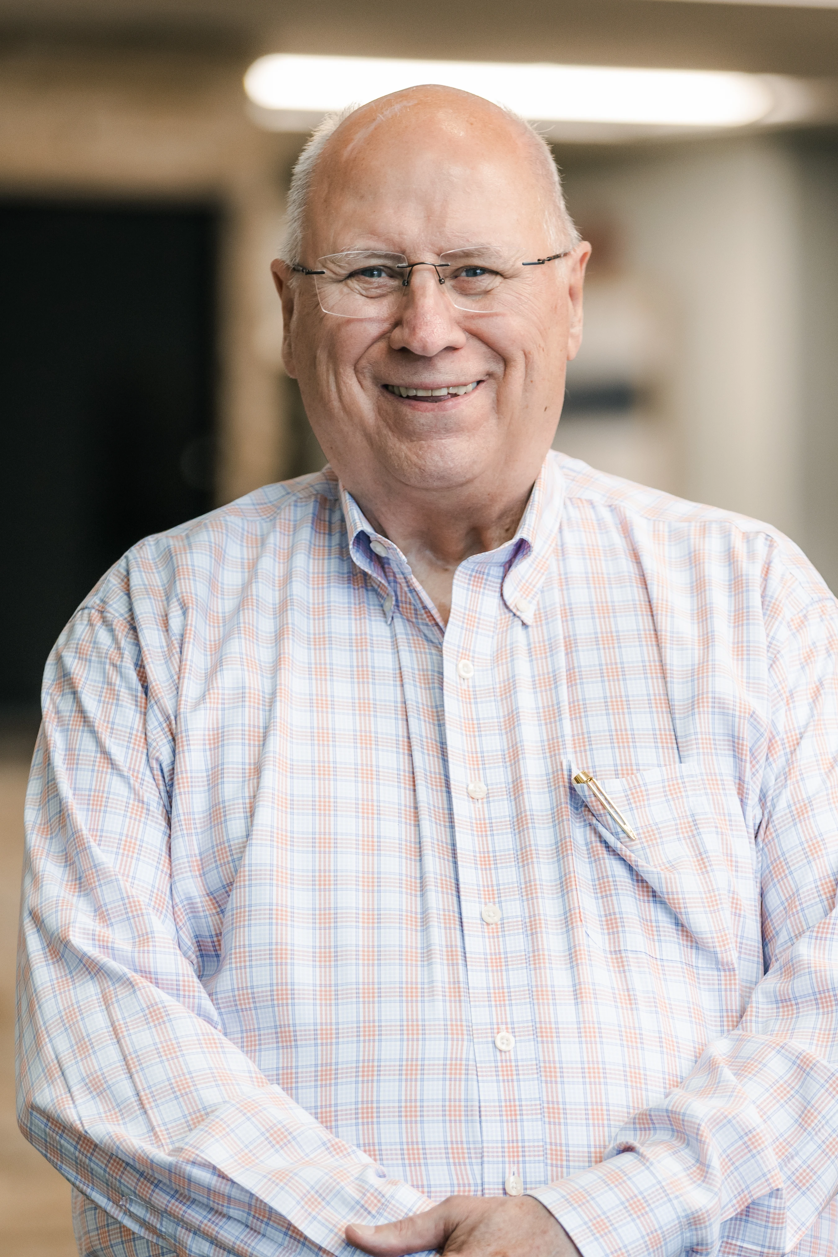 A friendly older man with glasses and a bald head smiles at the camera. He wears a light blue and orange plaid button-down shirt with a pen in his pocket.