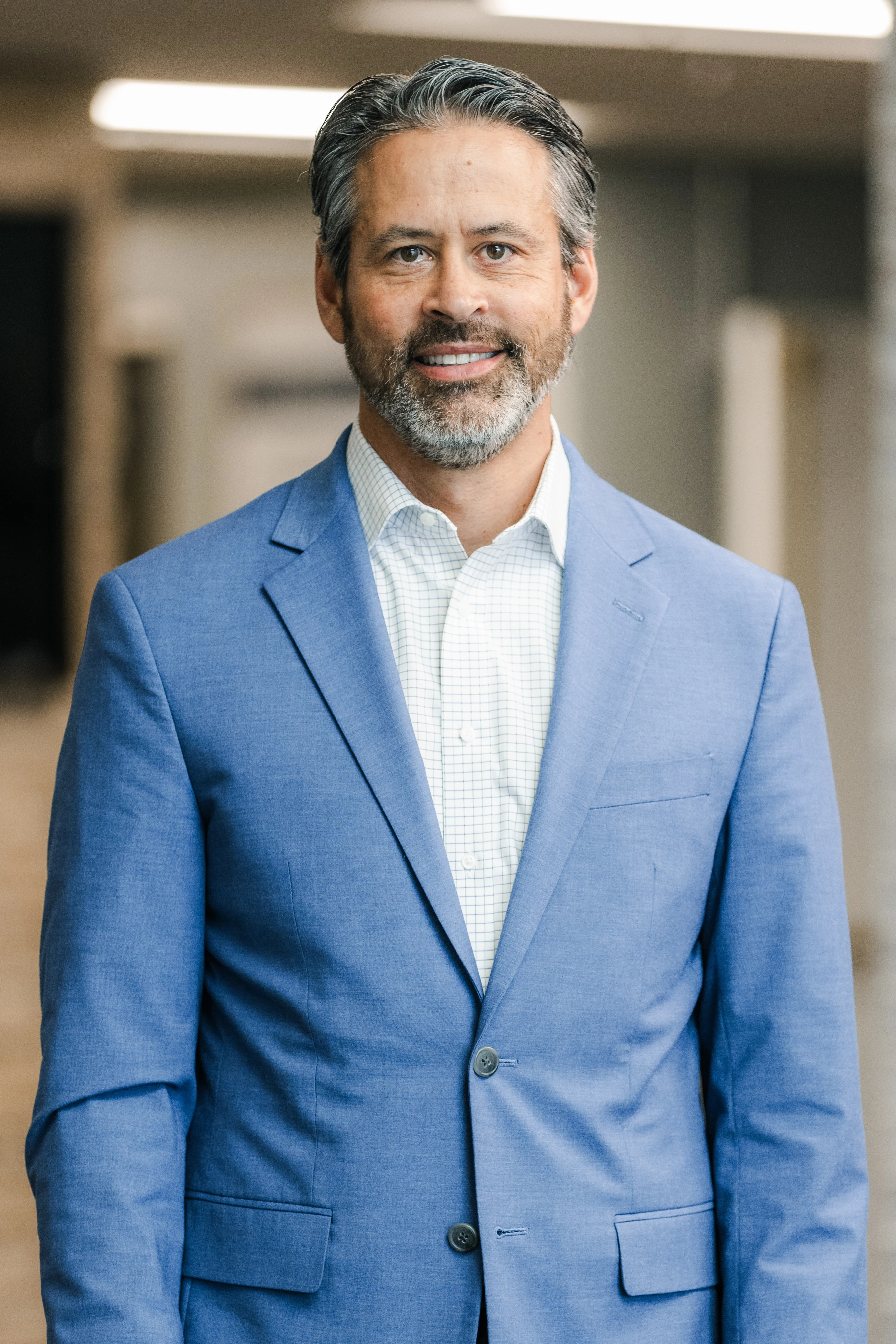 A smiling man with salt-and-pepper hair and beard poses for a headshot in a blue blazer and checkered shirt, standing in a brightly lit hallway.