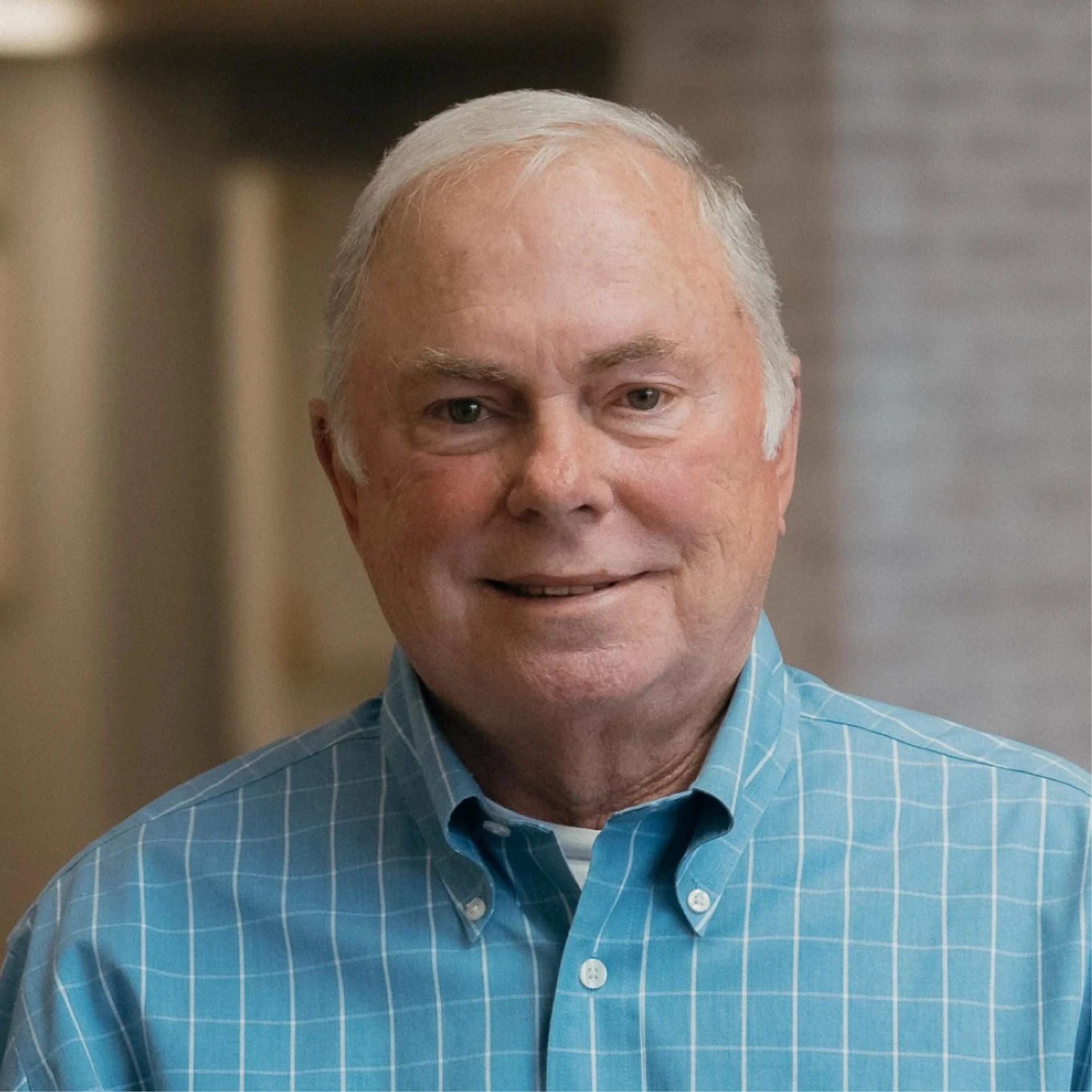 A headshot of an older man with short white hair and a gentle smile, wearing a light blue button-down shirt with a white grid pattern.