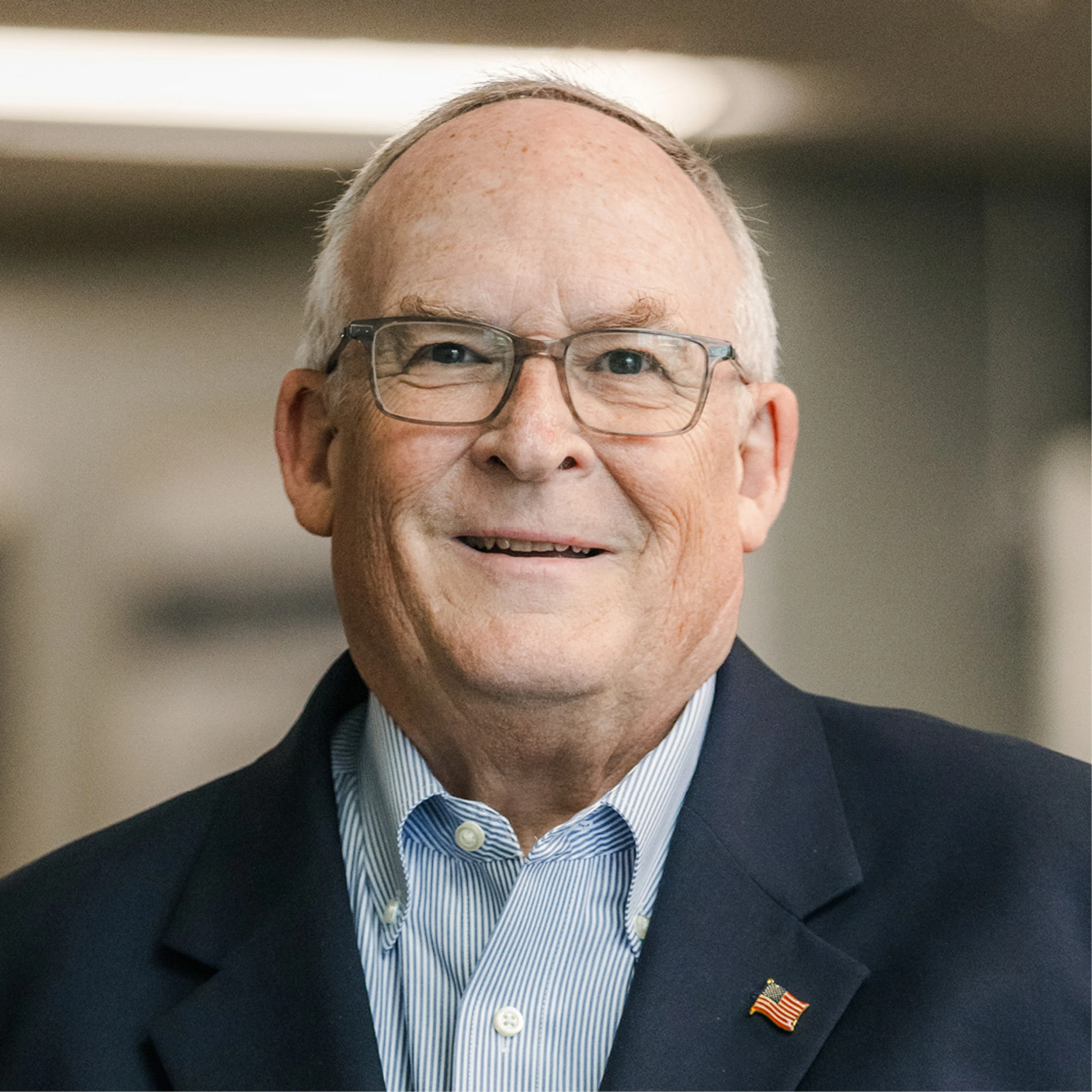 A head and shoulders portrait of a smiling older white man with glasses. He is wearing a blue striped shirt, a dark blazer, and an American flag lapel pin.