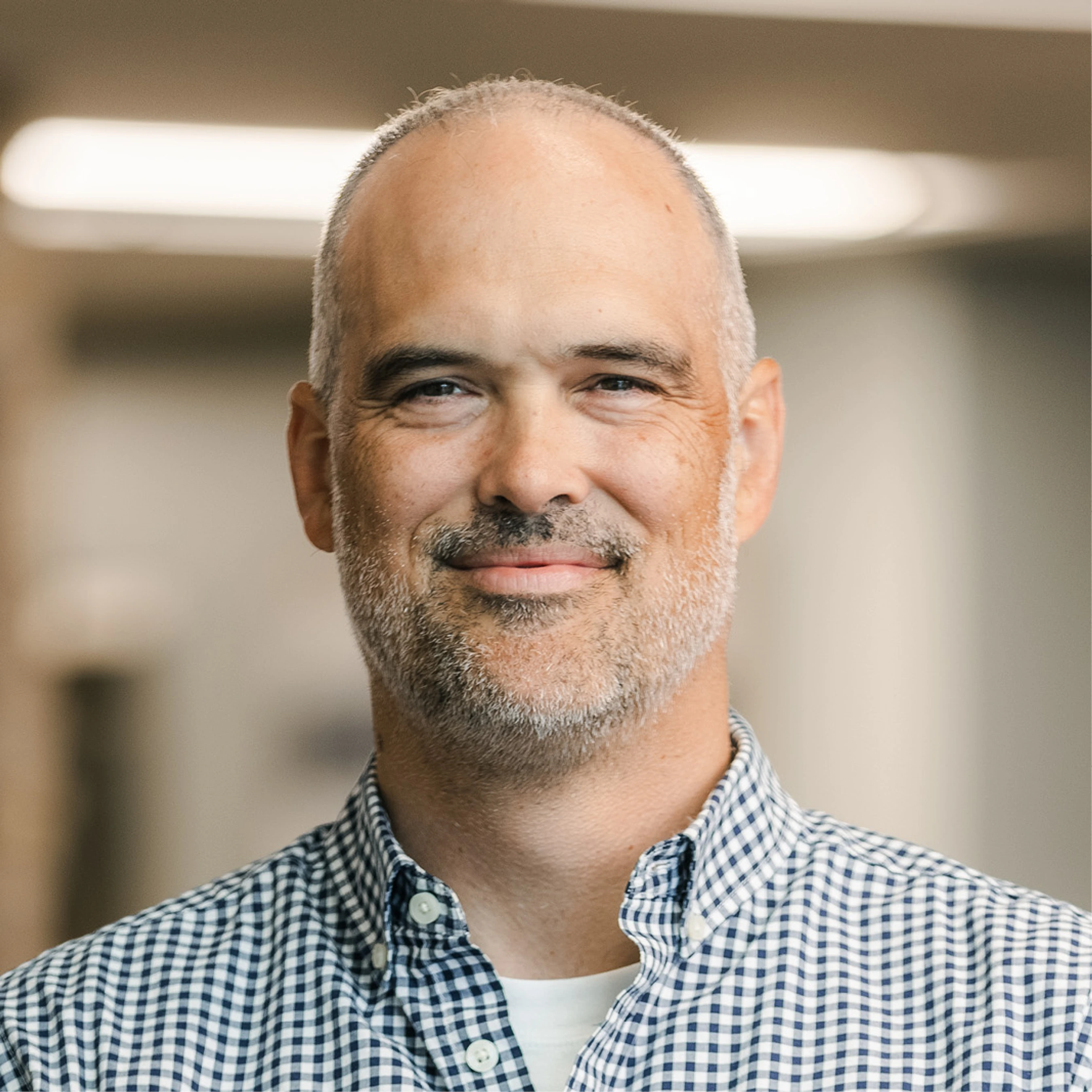 A headshot of a smiling middle-aged man with short gray hair and a beard, wearing a blue and white checkered button-down shirt in a softly lit hallway.