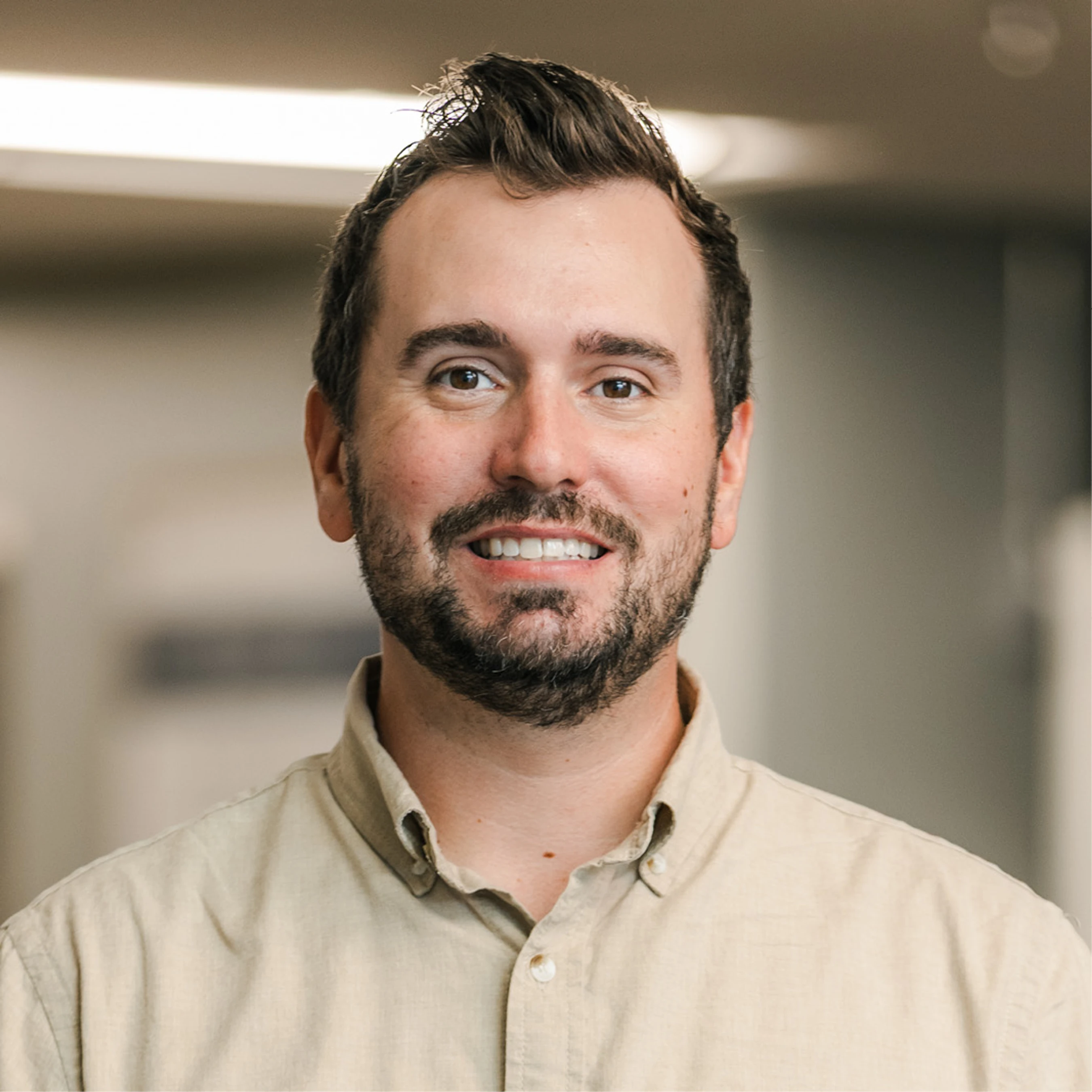 A headshot of a smiling man with short dark hair and a beard, wearing a beige collared shirt in a softly lit, out-of-focus office environment.