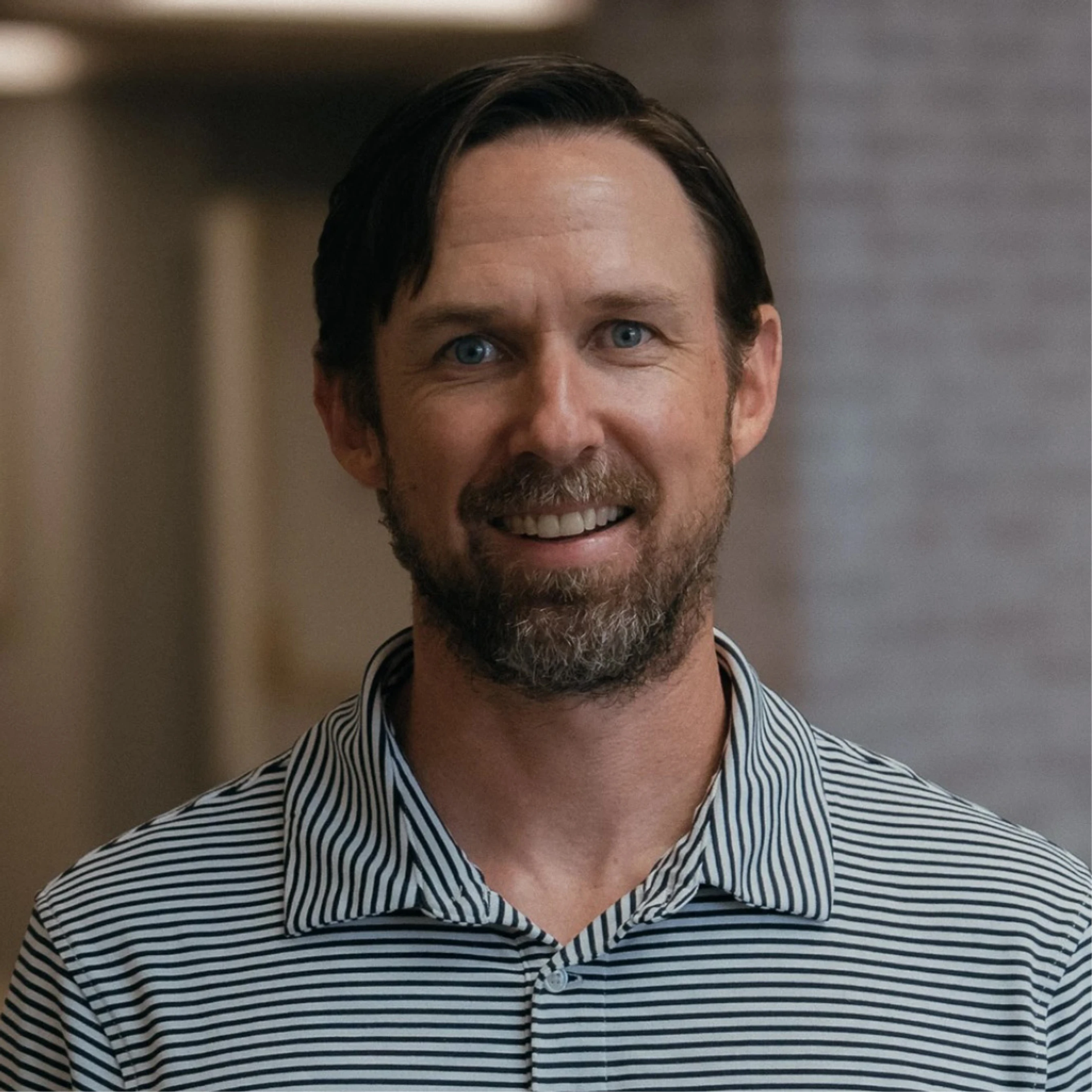 A head-and-shoulders portrait of a smiling middle-aged man with blue eyes, brown hair, and a beard, wearing a black and white striped polo shirt.
