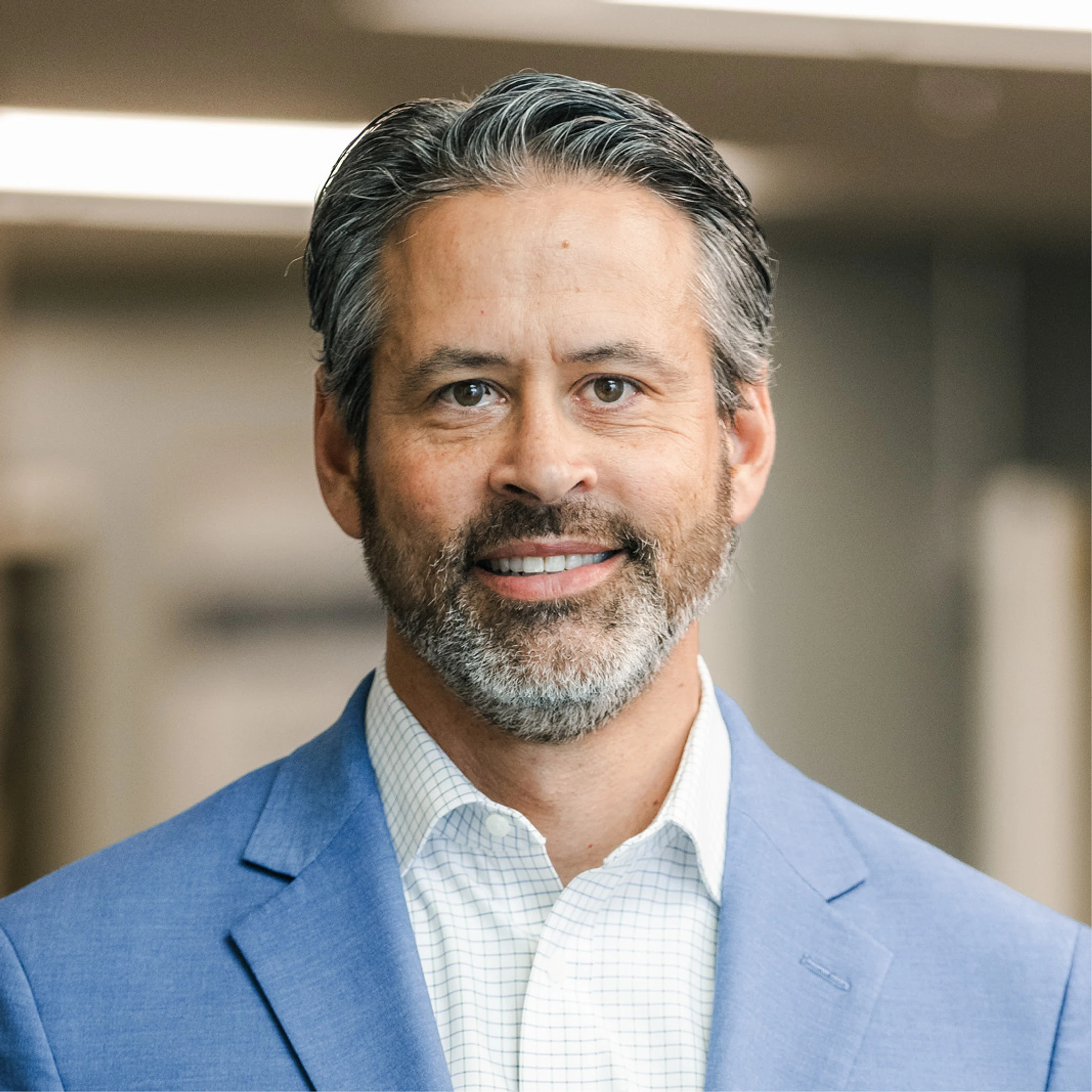 A head-and-shoulders portrait of a smiling, middle-aged man with salt-and-pepper hair and a beard, wearing a blue blazer and a checkered shirt.