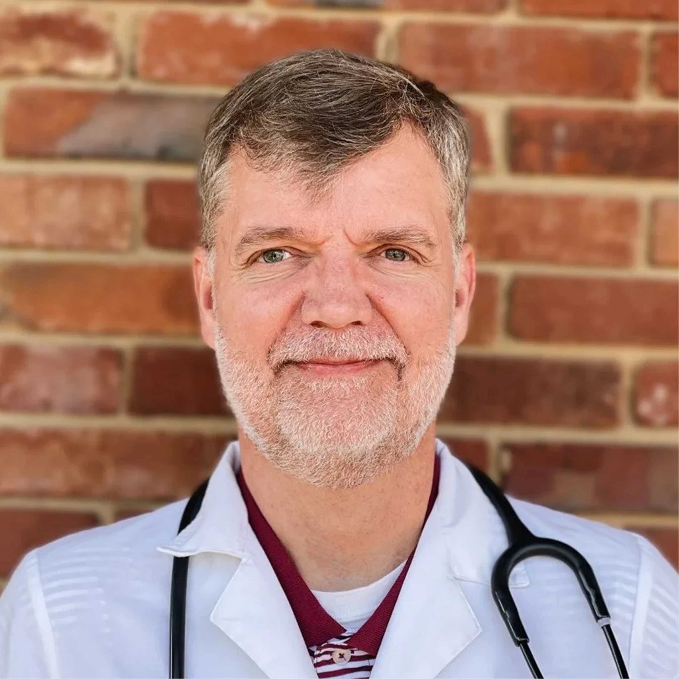 A headshot of a middle-aged male doctor with graying hair and a beard, wearing a white lab coat and a stethoscope, smiling in front of a brick wall.