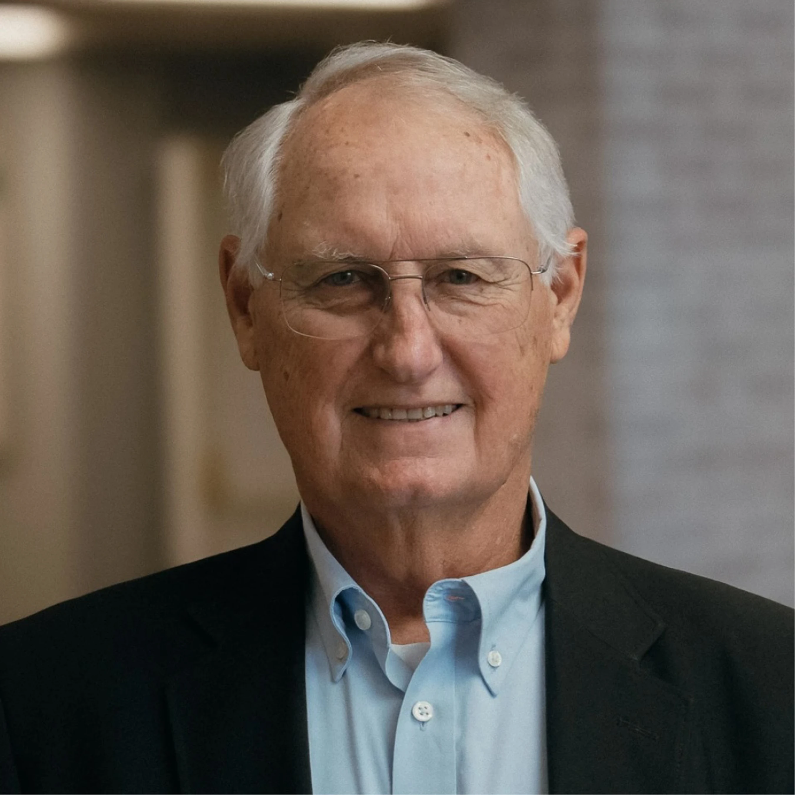 A head-and-shoulders portrait of an older man with white hair and glasses, smiling gently. He is wearing a light blue collared shirt and a dark blazer.