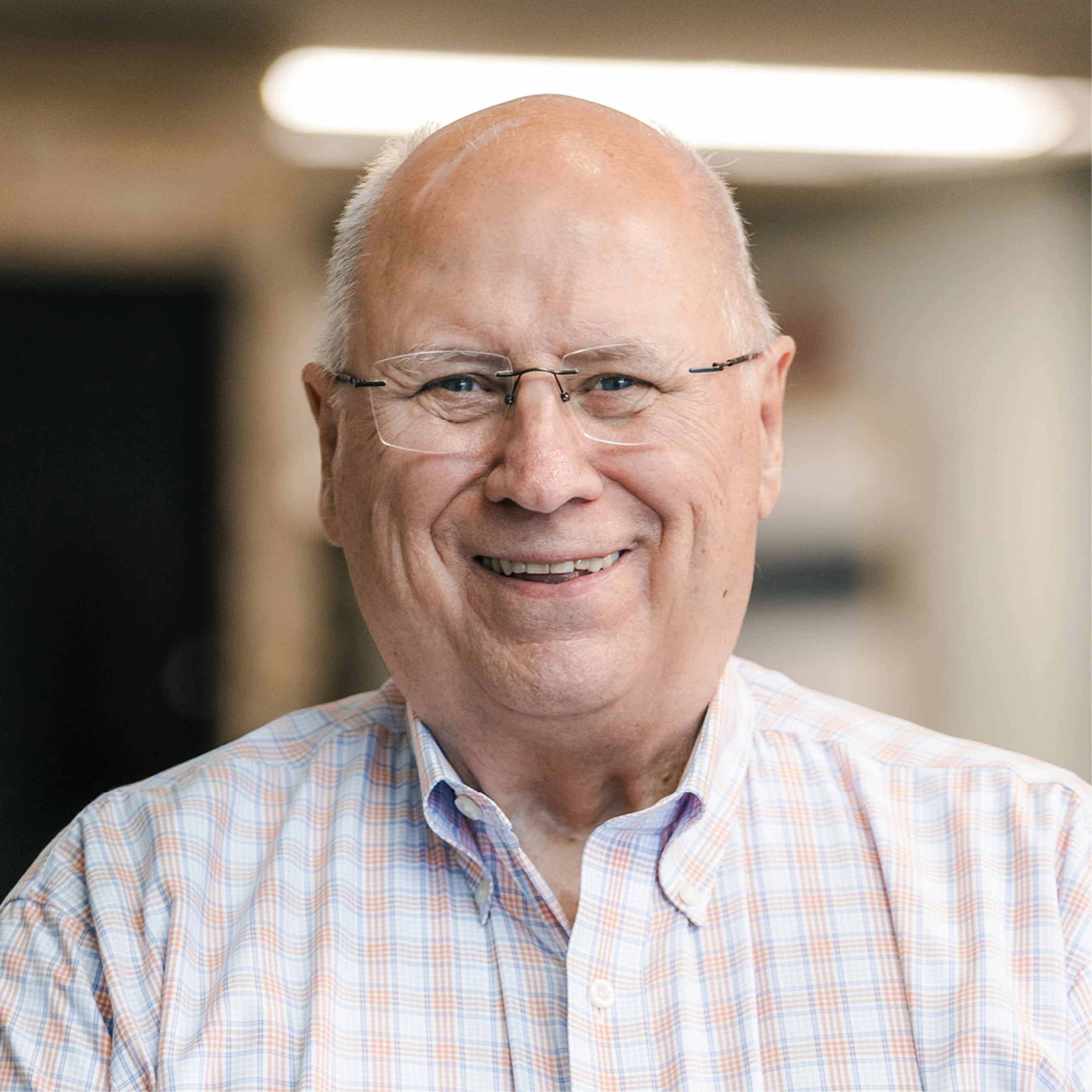 A friendly older white man with a bald head and glasses smiles for a headshot. He is wearing a light-colored plaid button-down shirt indoors.