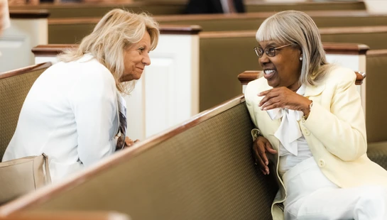 A white woman and a Black woman sit in church pews, smiling as they engage in a warm and friendly conversation with each other.