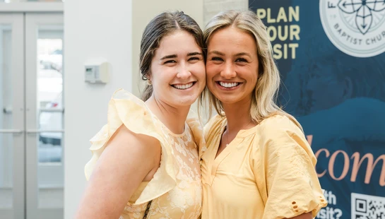 Two young women in coordinating yellow dresses stand close together, smiling warmly at the camera inside a church lobby next to a welcome banner.