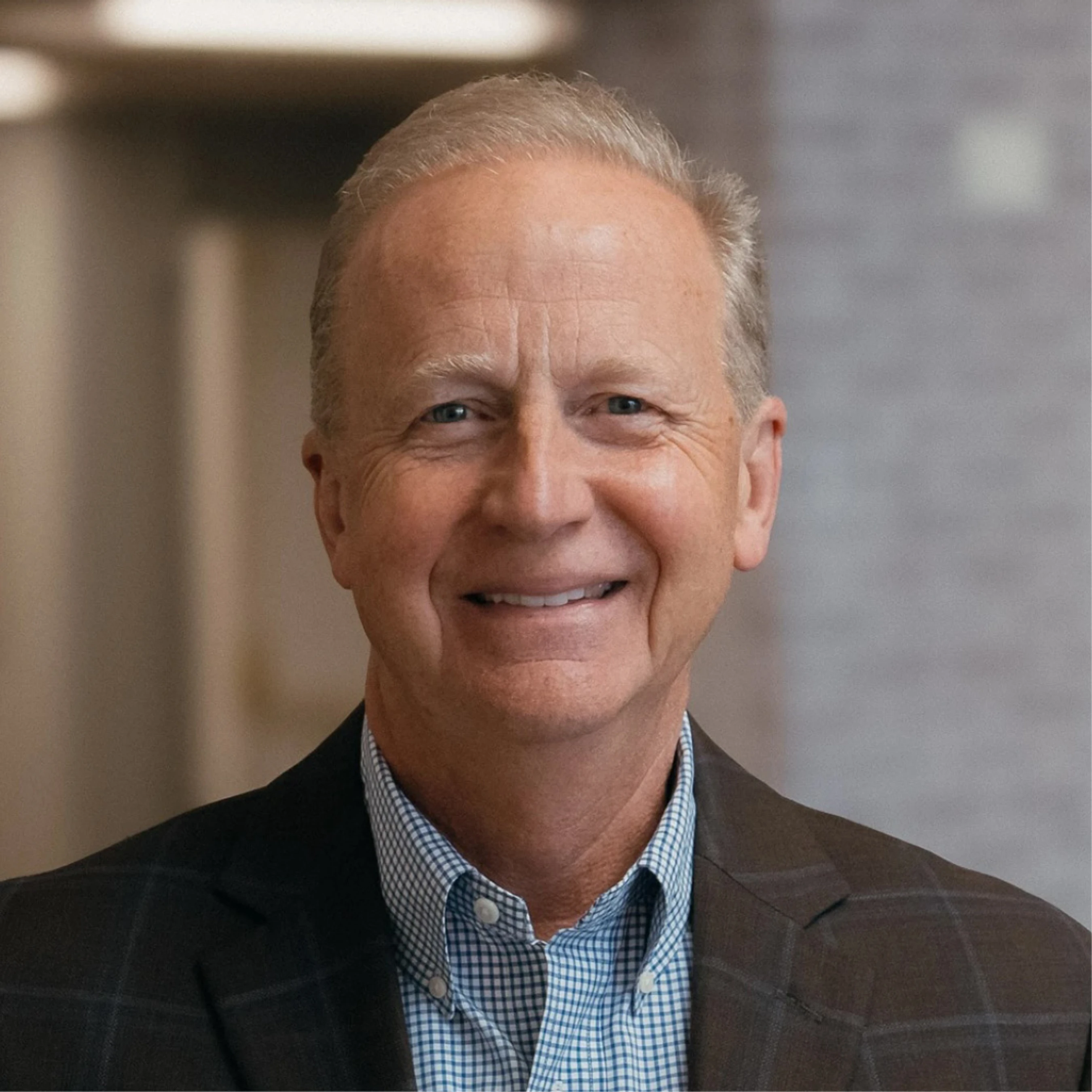 A headshot of an older white man with gray hair, smiling at the camera. He is wearing a blue checkered shirt and a dark plaid blazer in an office.