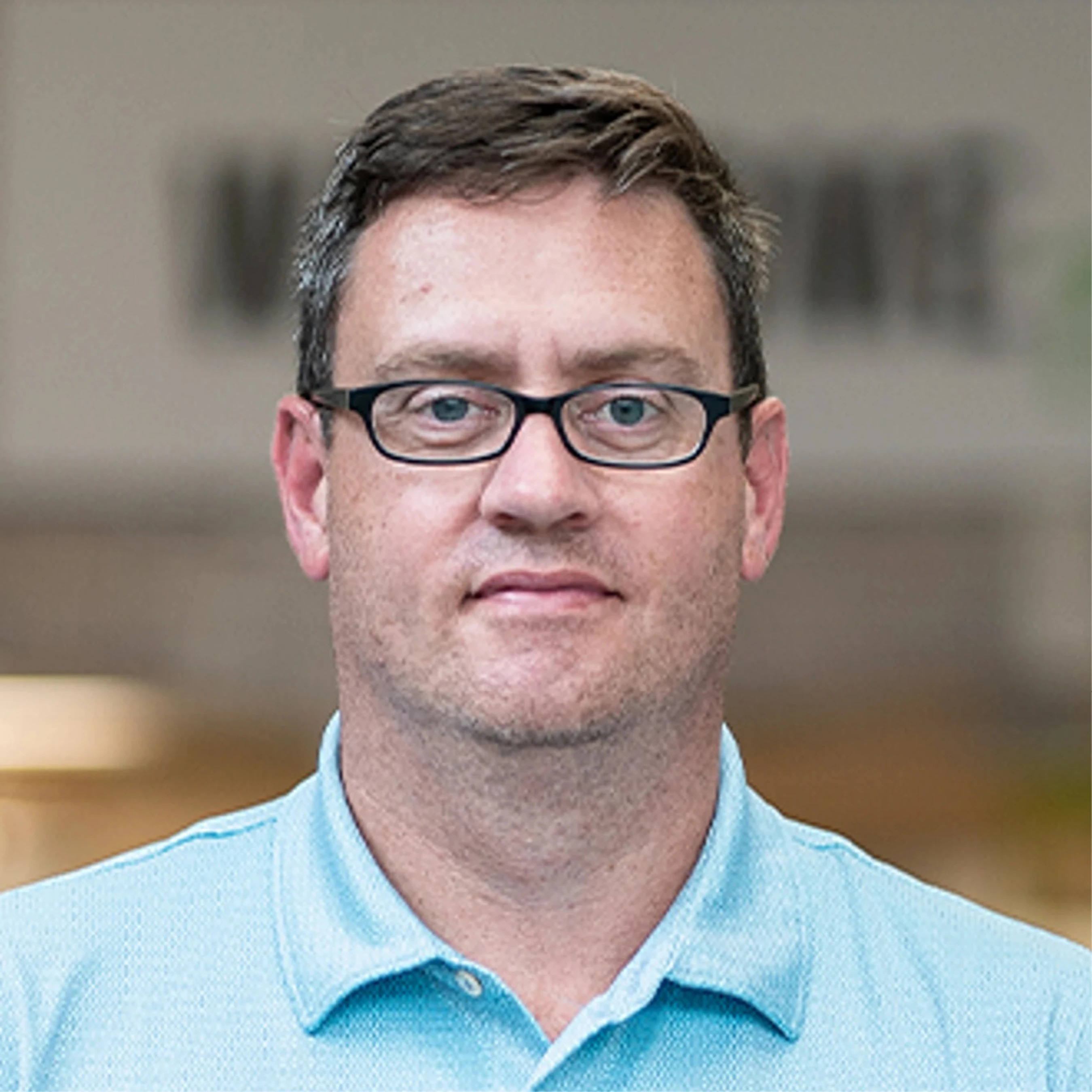 A head-and-shoulders portrait of a middle-aged man with short, dark hair and black-framed glasses, wearing a light blue polo shirt against a blurred background.