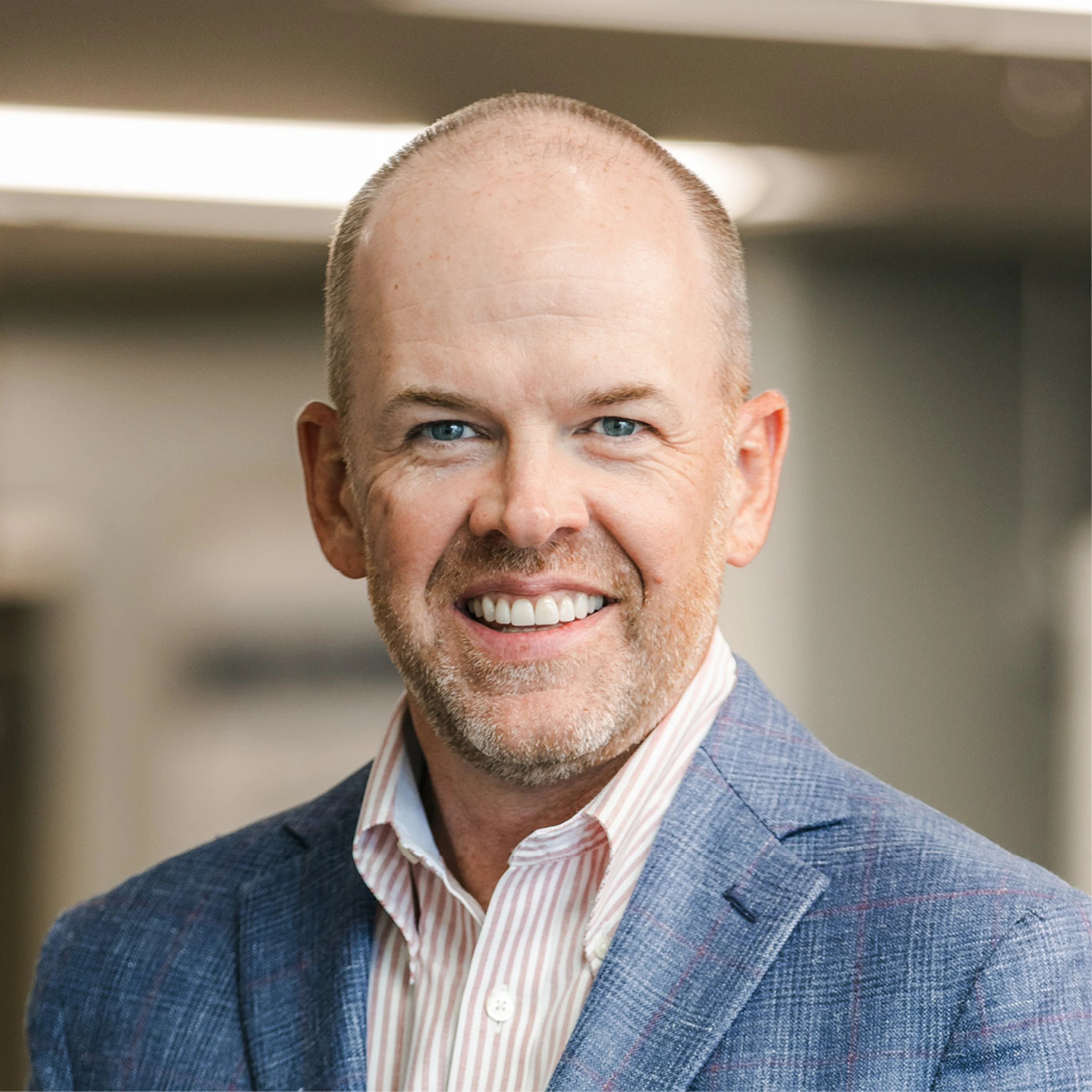 A friendly, smiling, middle-aged man with blue eyes and short-cropped hair poses for a headshot in a blue blazer and striped collared shirt.