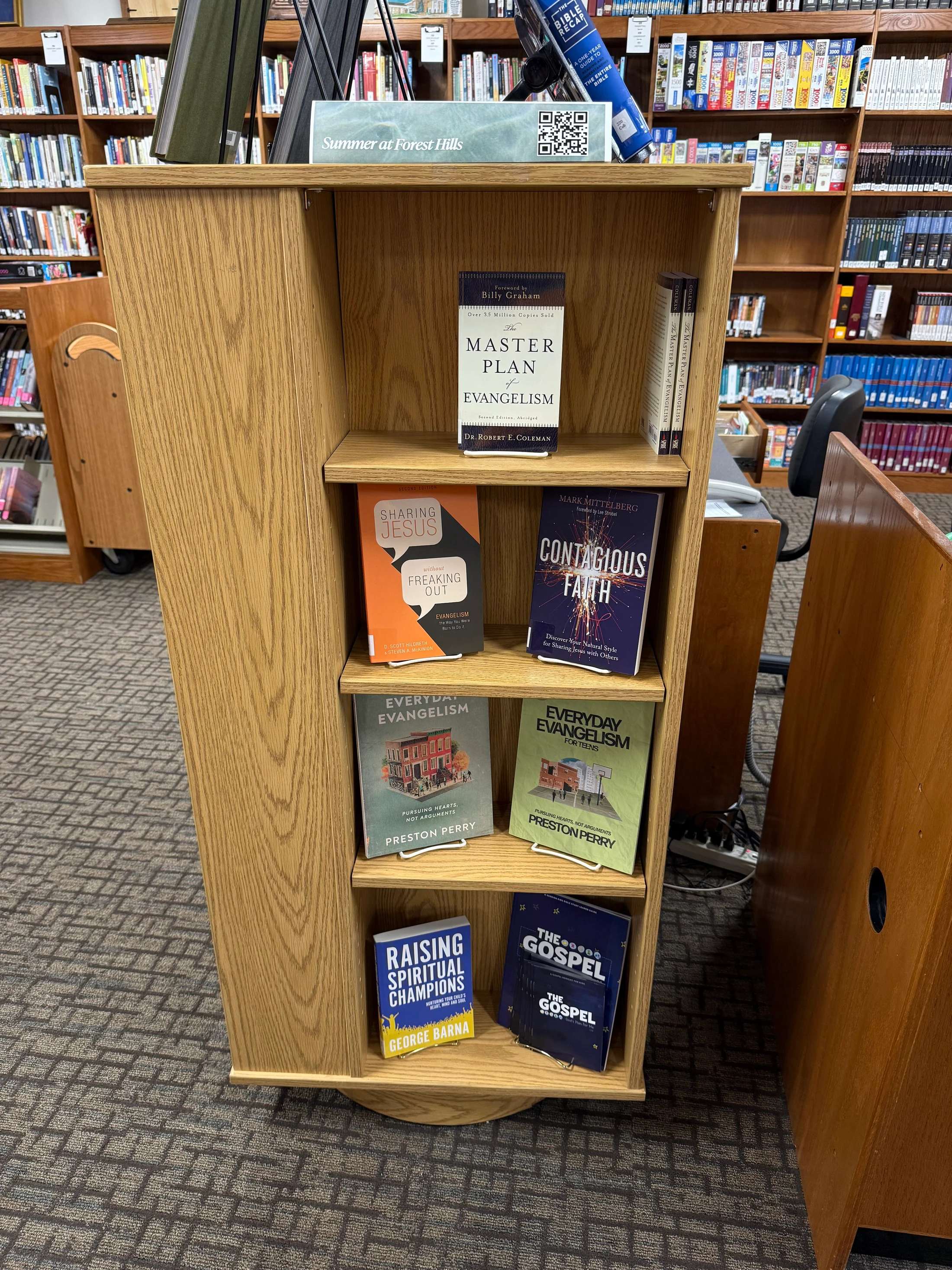 A wooden rotating bookshelf in a library displays a collection of Christian books about evangelism, with other bookshelves visible in the background.