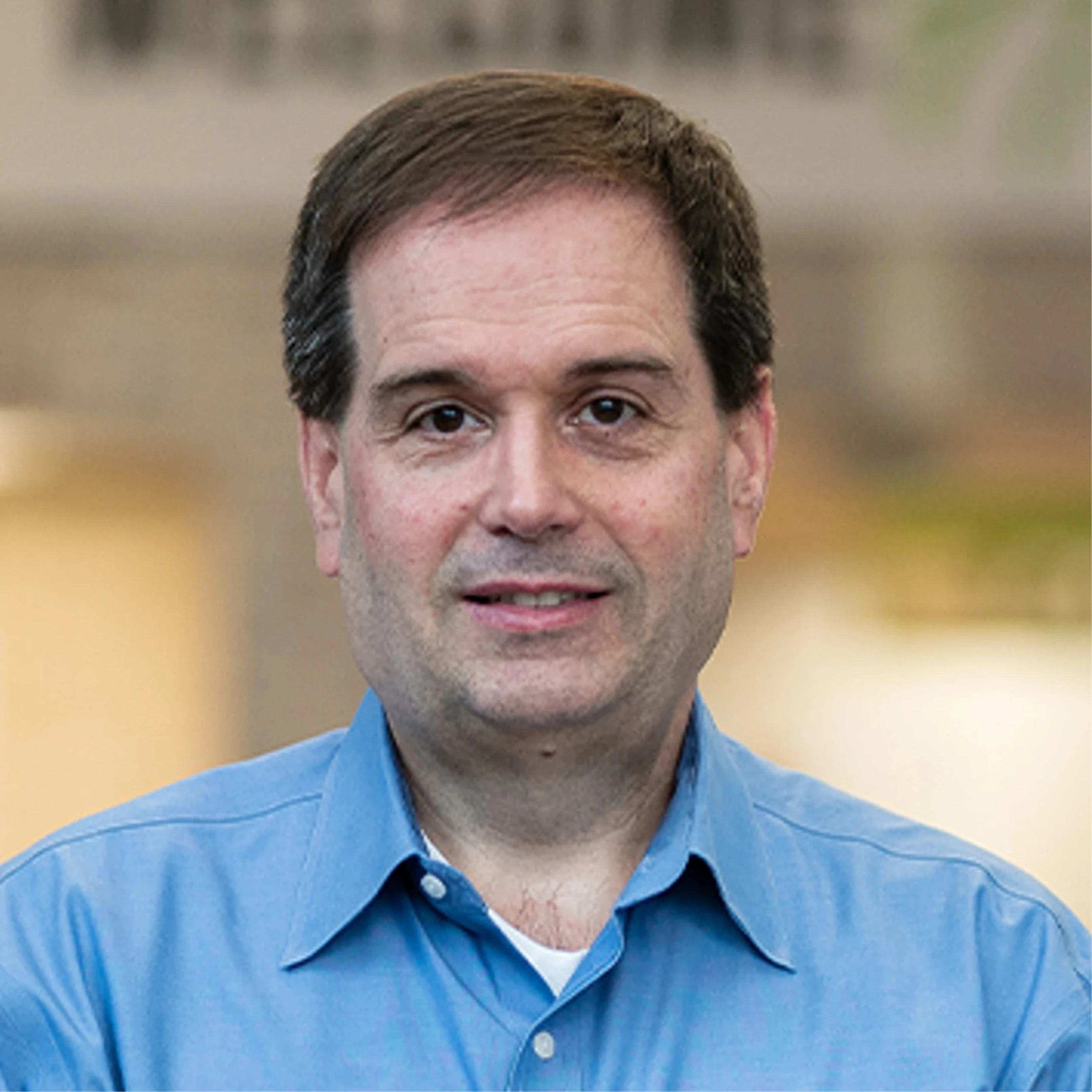 A professional headshot of a middle-aged man with dark hair and brown eyes, wearing a blue button-down shirt and smiling at the camera.